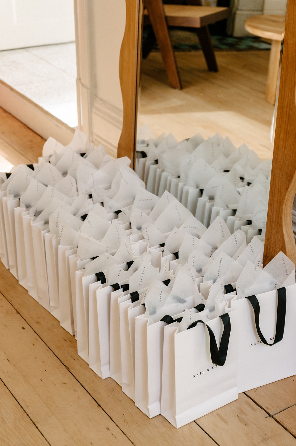Elegant white gift bags with black handles filled with tissue paper, neatly arranged on a wooden floor, reflecting in a large mirror.