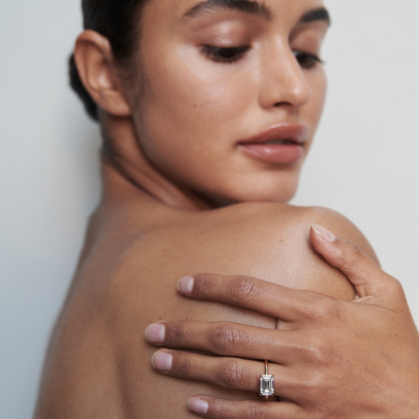 Woman showcasing a luxurious, emerald-cut diamond ring on her finger, against her bare shoulder. Elegant and minimalist jewellery display.