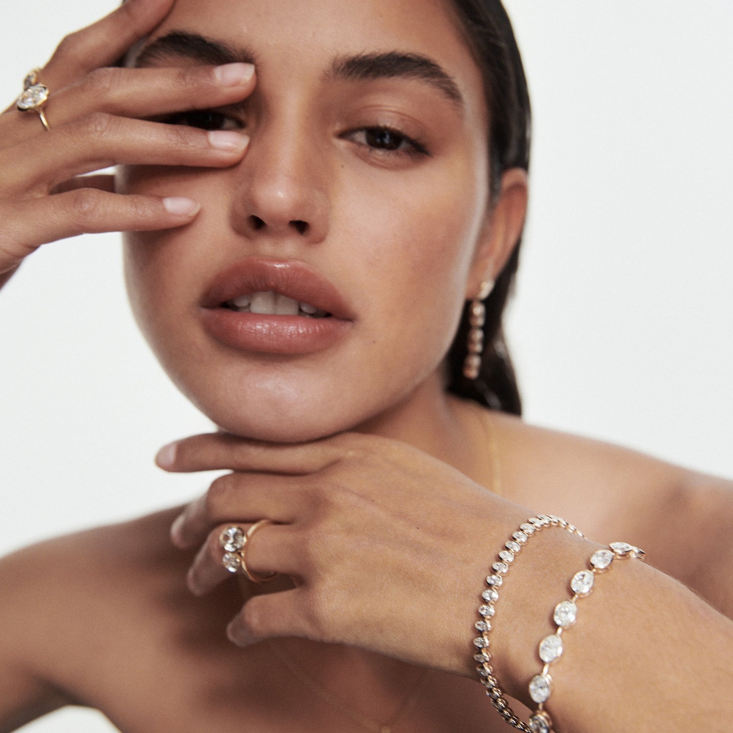 Woman wearing luxury jewellery including diamond bracelets, earrings, and rings, posed with hand near face against a soft-focus background.