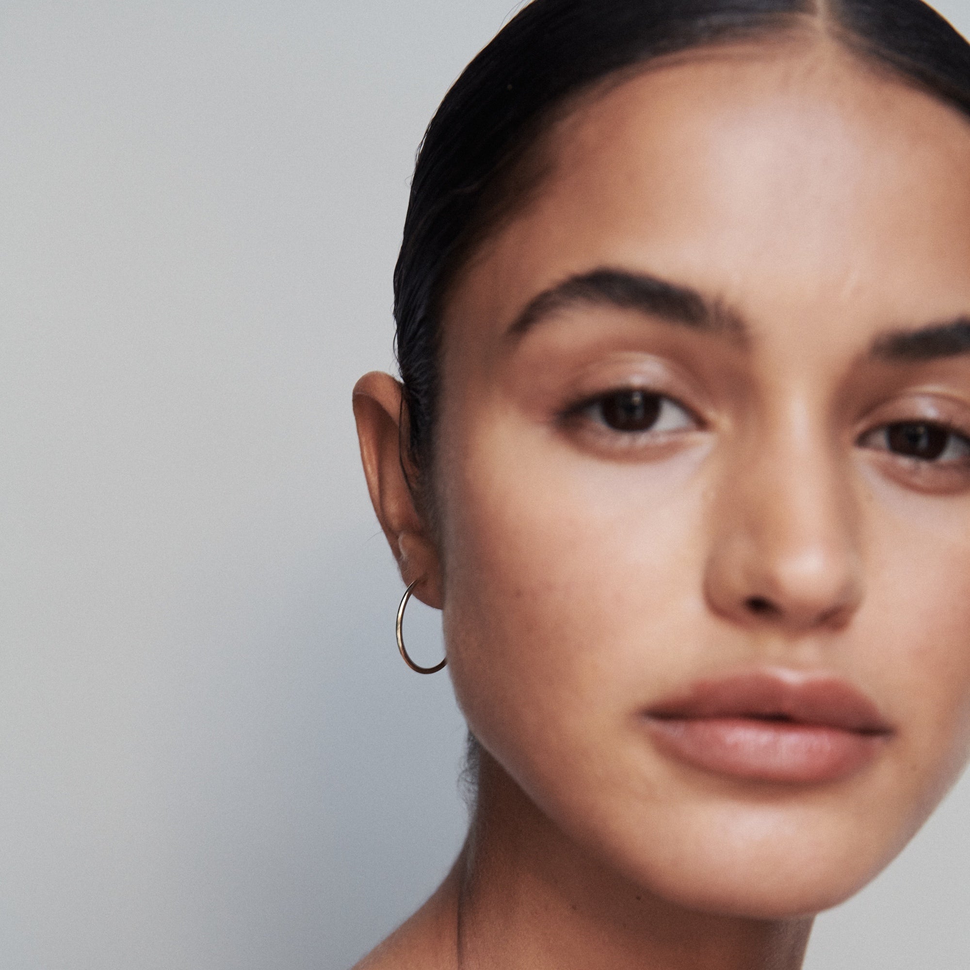 Woman wearing a sleek gold hoop earring, posing with a neutral expression against a plain background. Luxury jewellery focus.