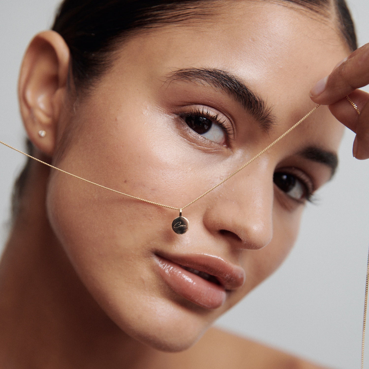 Woman holding a delicate gold necklace with a small round pendant near her face. Minimalist luxury jewellery, portrait style.