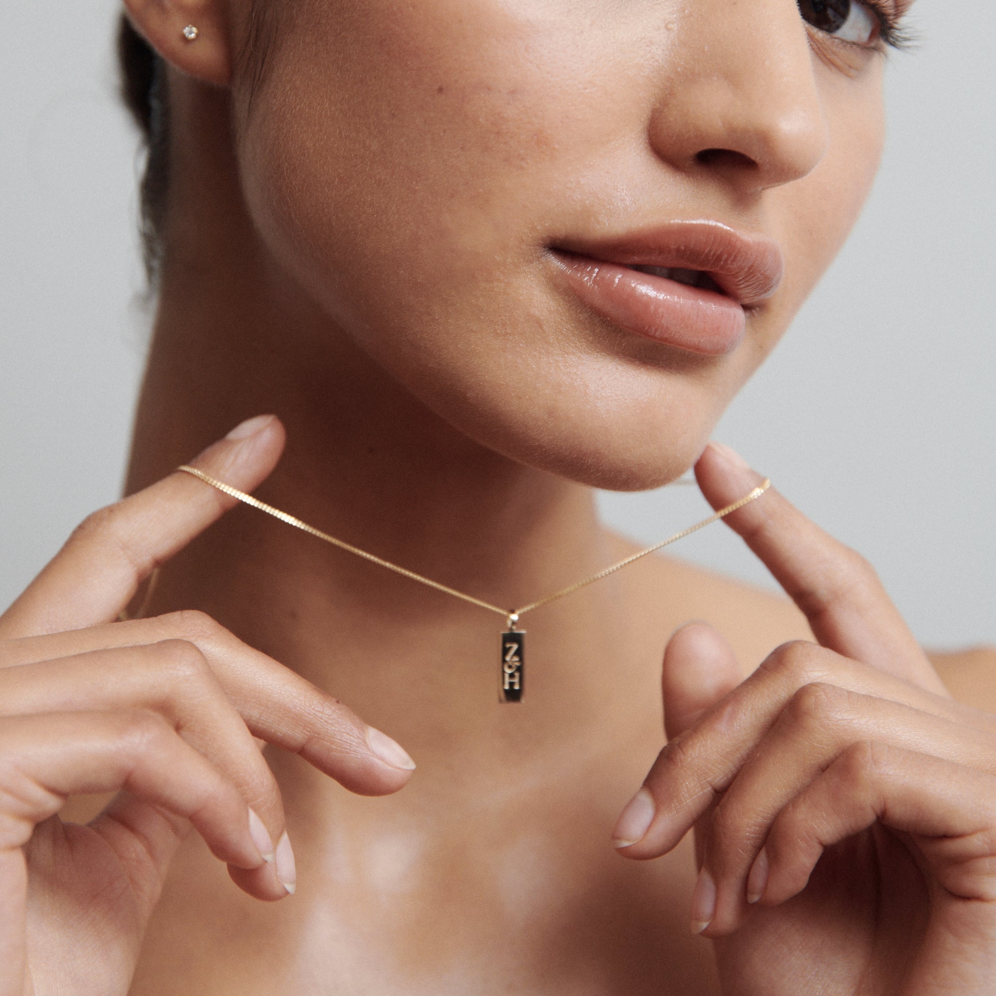 Woman wearing a minimalist gold necklace with a vertical pendant, showcasing high-end jewellery. Close-up of her face and fingers touching the chain.