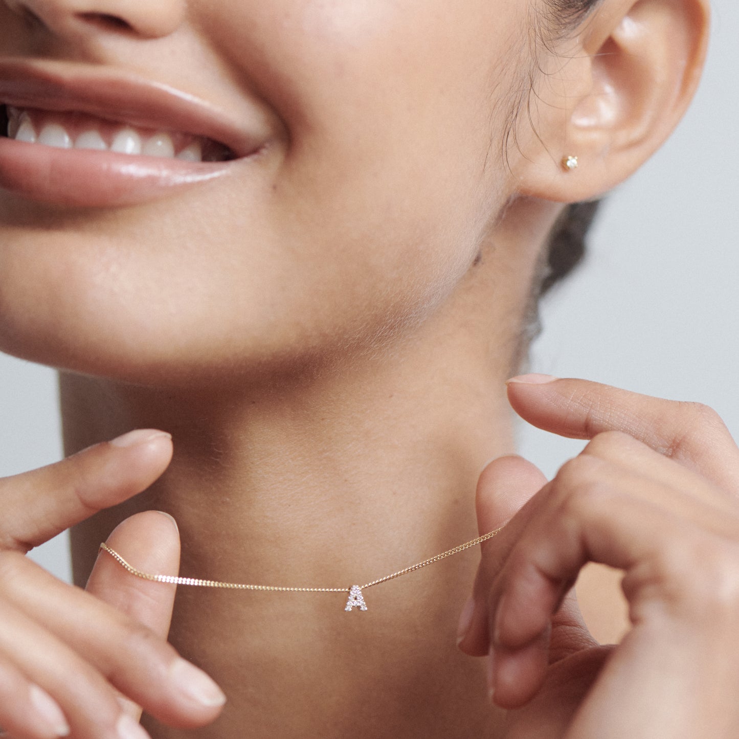 Woman smiling, showcasing a delicate gold chain necklace with an 'A' pendant, complemented by a small stud earring. Luxurious jewellery focus.