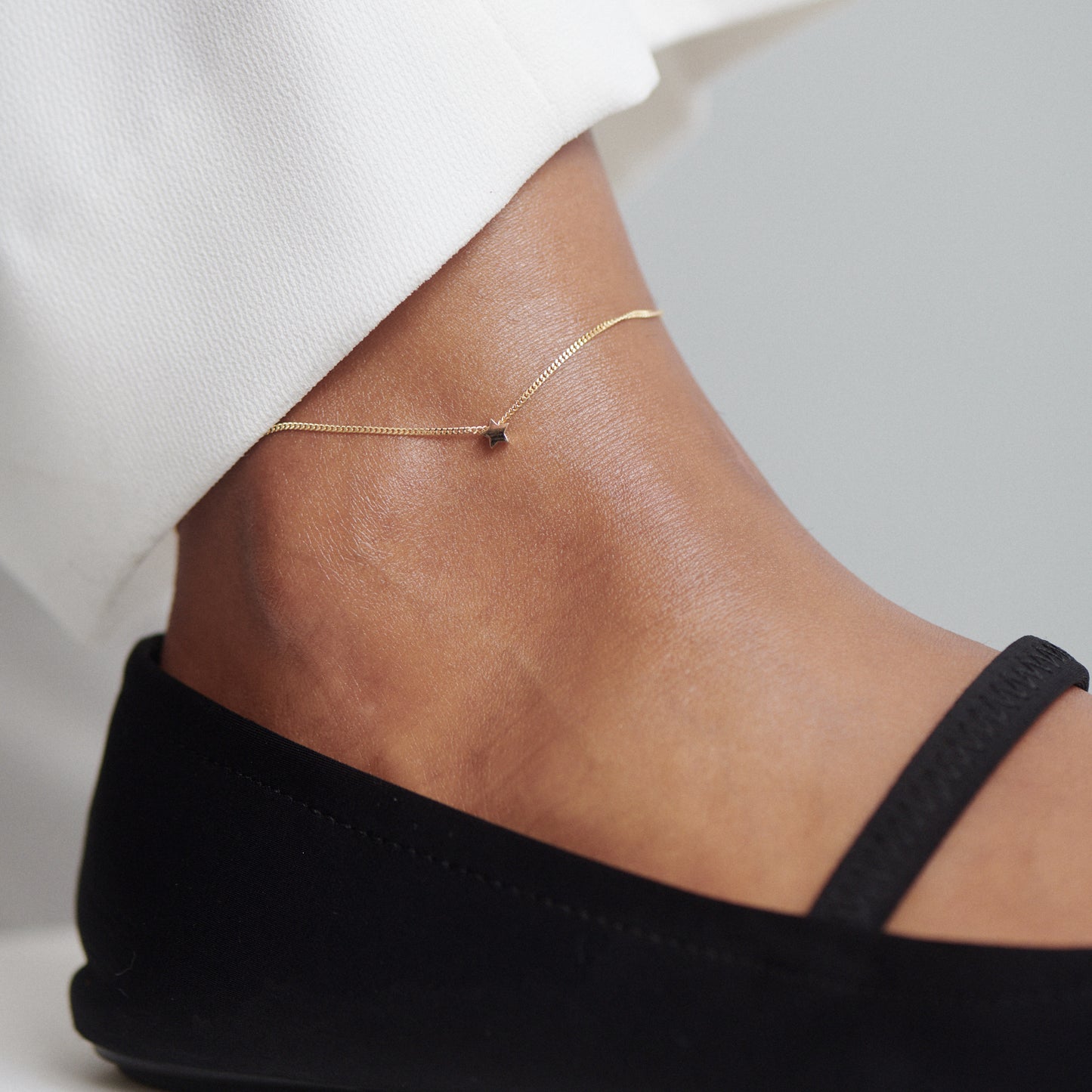 Gold anklet with delicate charm on ankle, paired with white trousers and black flats. Luxurious minimalist jewellery style.