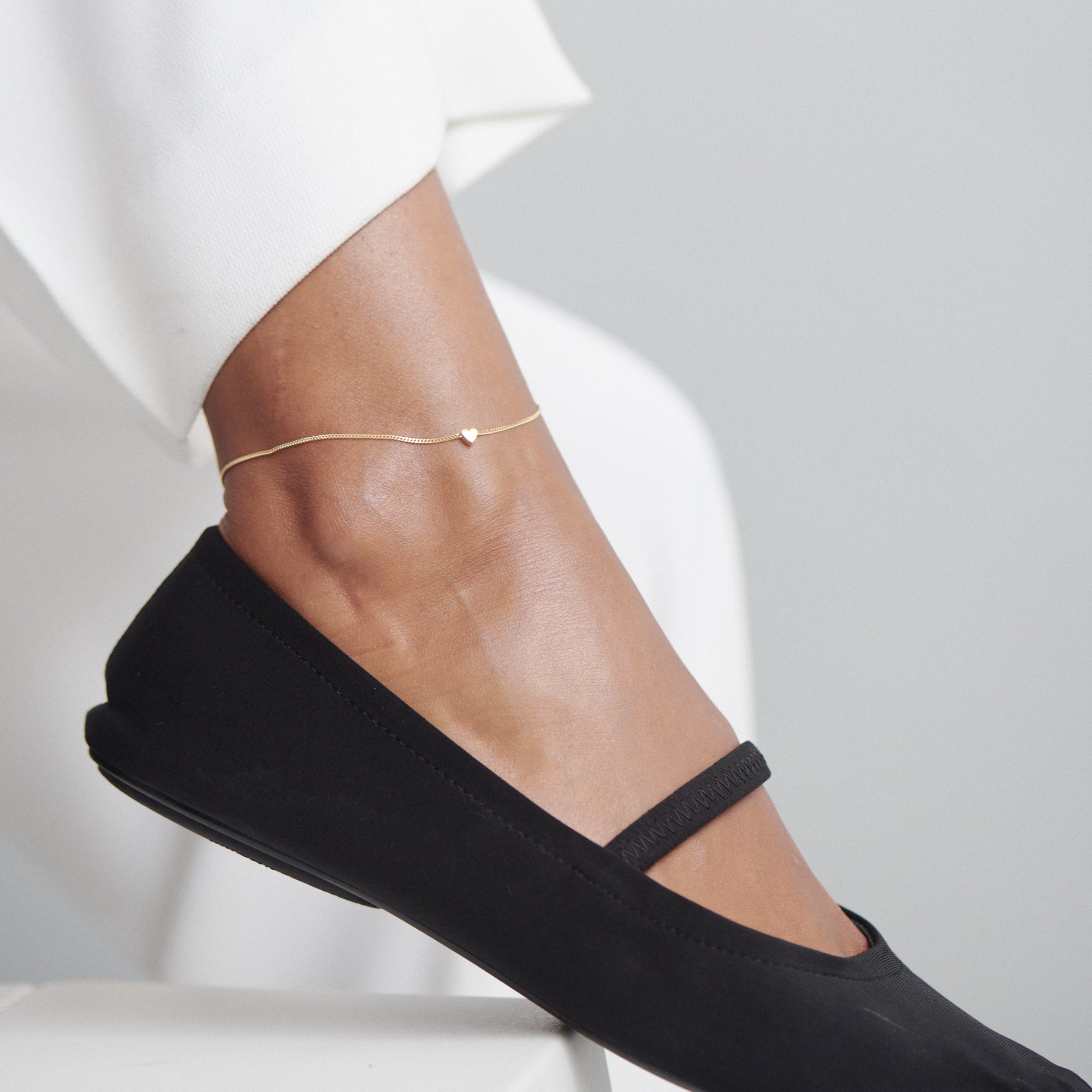 Gold anklet with a small heart charm on a leg wearing sleek black flats and white trousers; minimalist luxury jewellery.