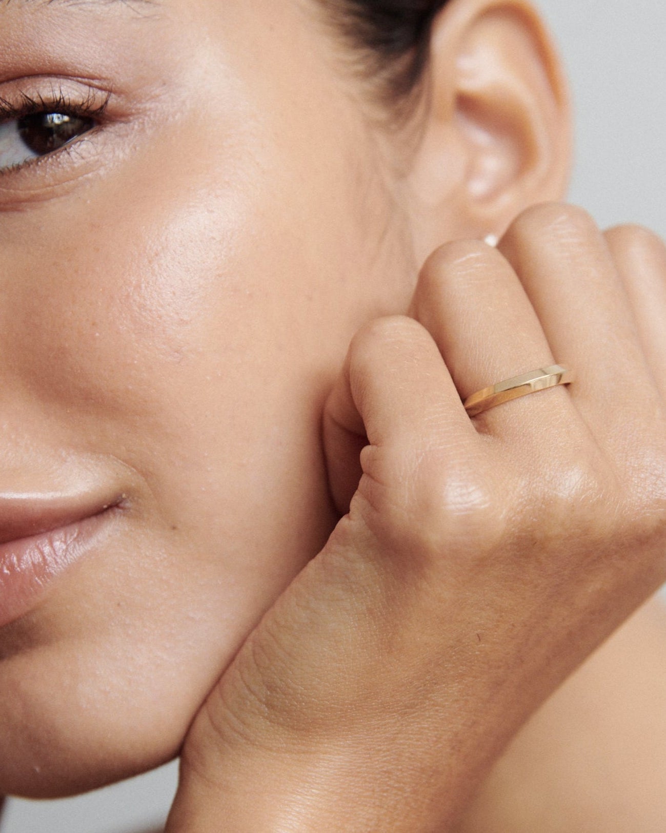 Woman's hand gracefully resting by face, showcasing a sleek gold ring. Elegant high-end jewellery captured in soft light.