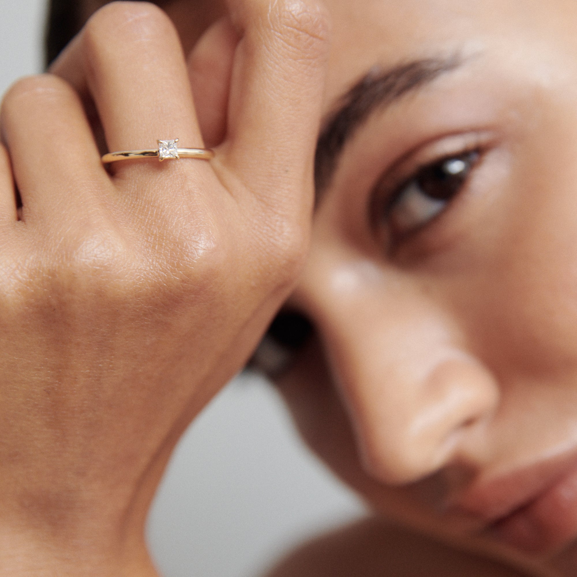 Close-up of a person wearing a gold ring with a square diamond, hand elegantly placed near their face. Luxury jewellery detail.