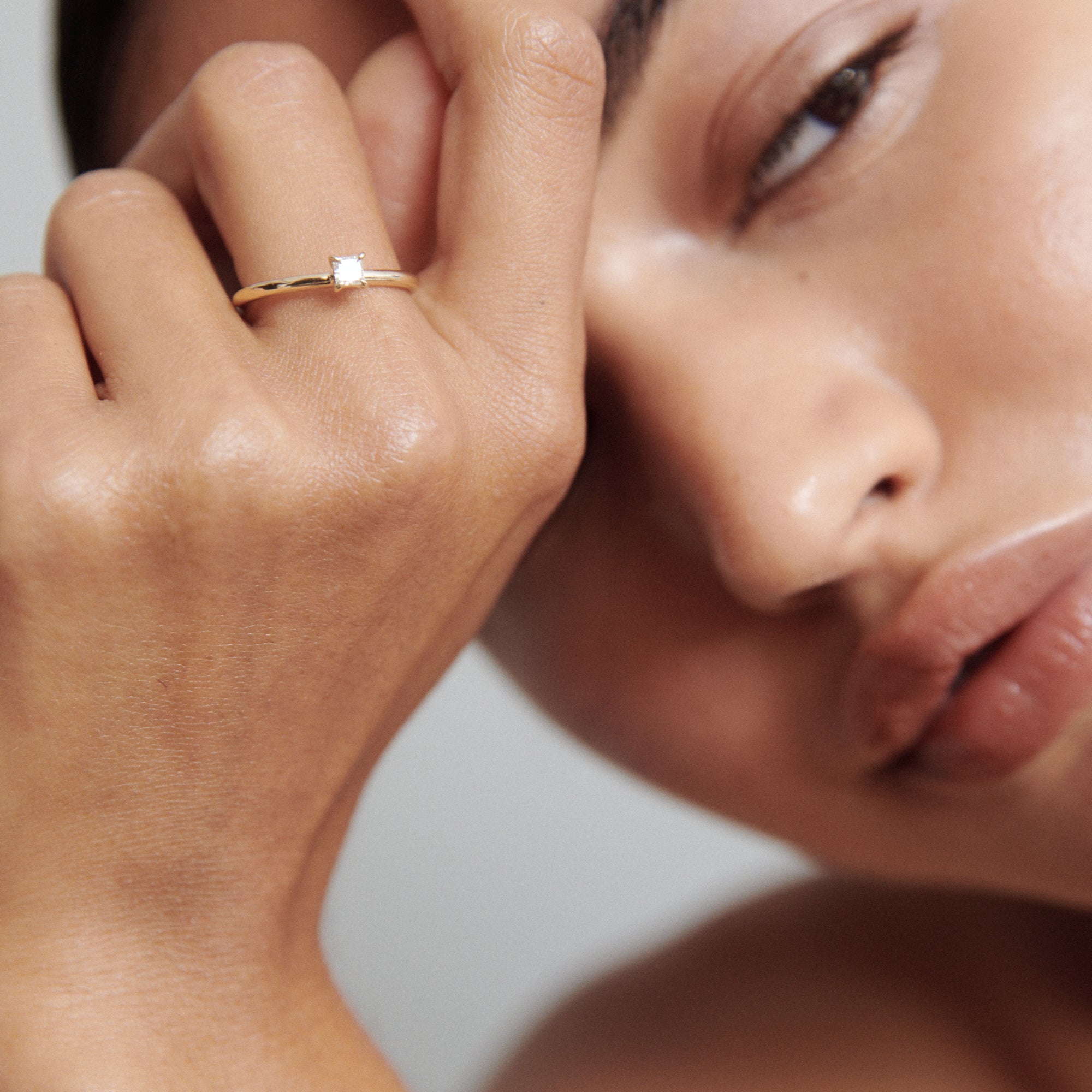 Woman's hand with a delicate gold solitaire ring near her face, showcasing luxury jewellery with a minimalist, elegant design.