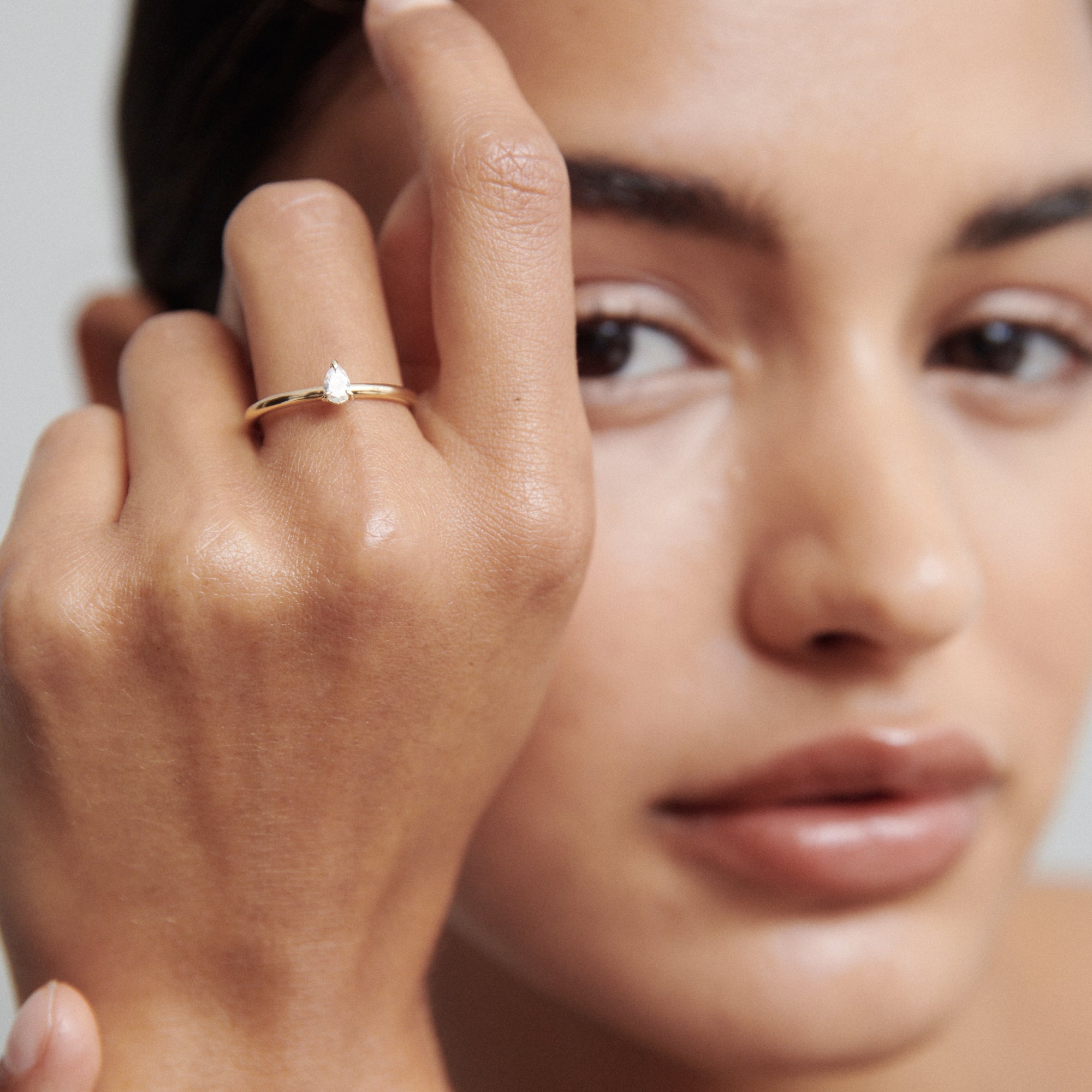 Close-up of a woman's hand with a gold ring featuring a small pear-shaped diamond. The woman's face is slightly blurred in the background.