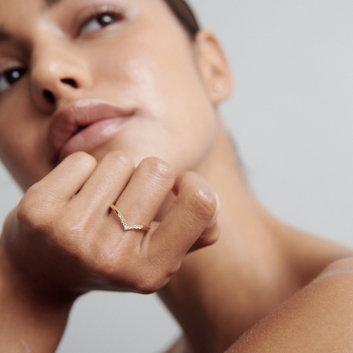 Woman's hand with delicate gold ring featuring small diamonds. Soft focus on face, neutral background. Luxury jewellery showcase.