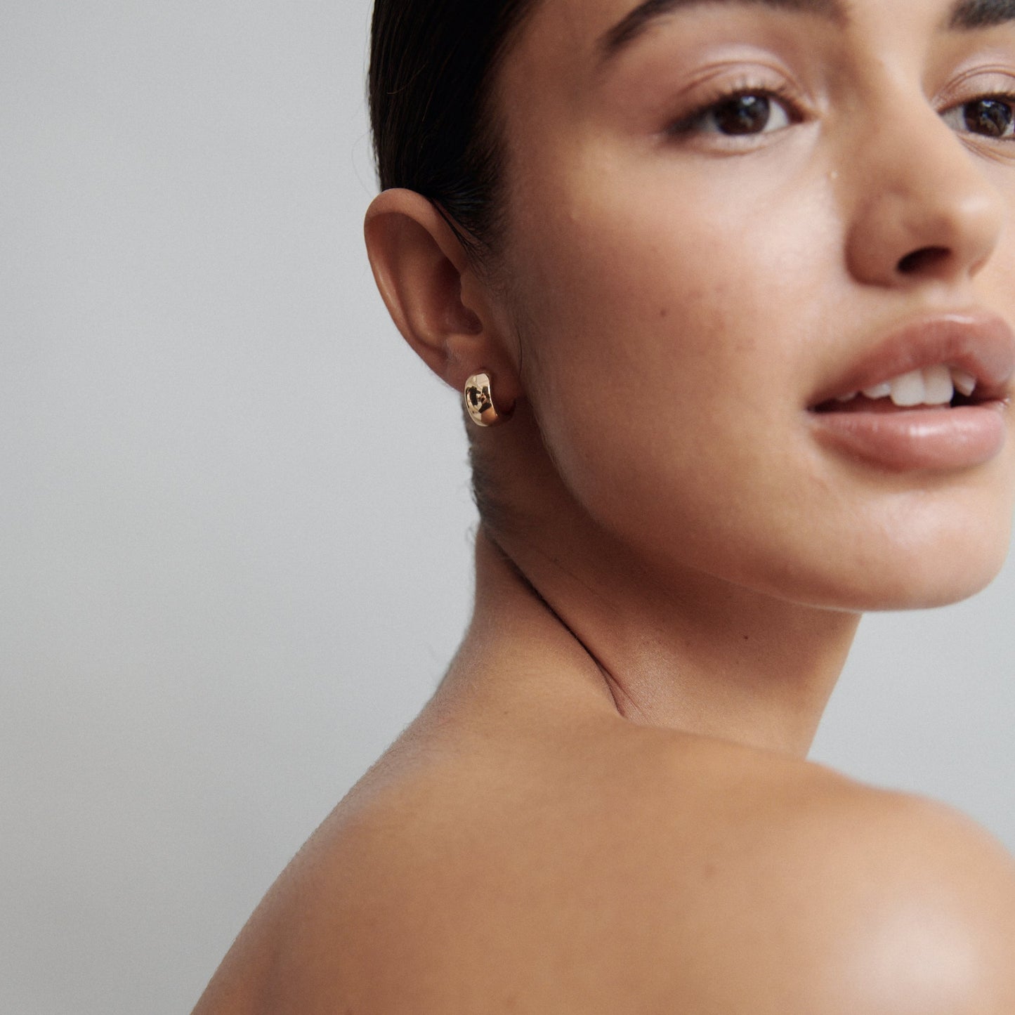 Woman wearing elegant gold hoop earring, with smooth skin and neutral makeup, photographed from the side against a light grey background.
