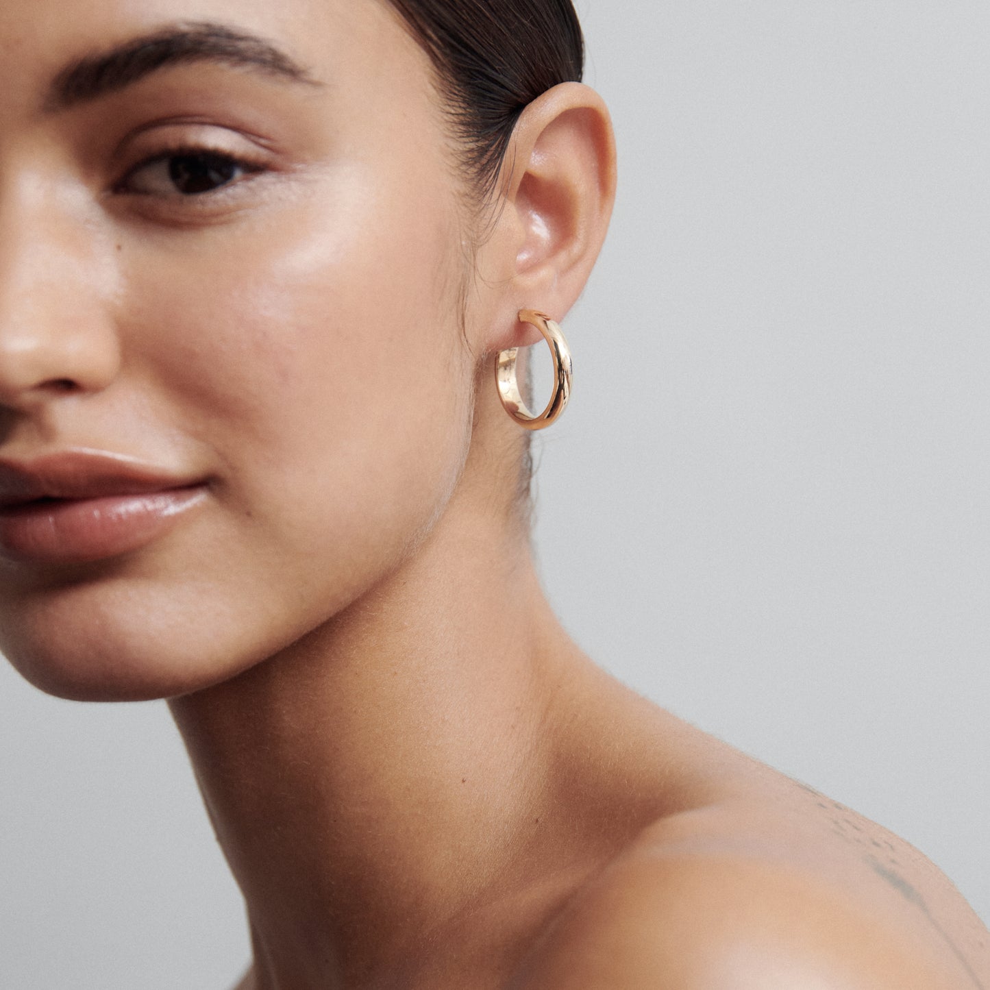 Close-up of a woman wearing a smooth gold hoop earring, showcasing minimalist luxury jewellery against a plain grey background.