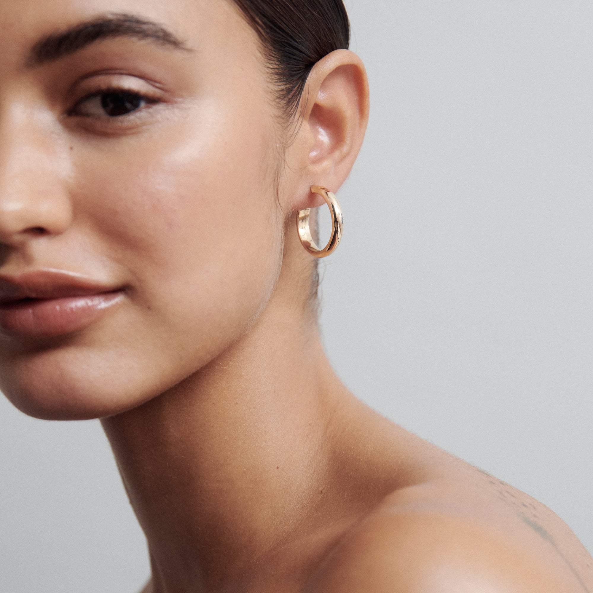 Close-up of a woman wearing a smooth gold hoop earring, showcasing minimalist luxury jewellery against a plain grey background.