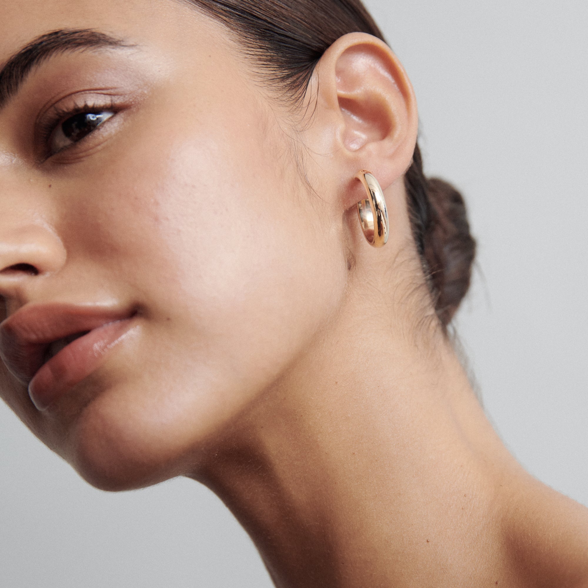 Woman wearing elegant gold hoop earrings, close-up profile. Hair is tied back, showcasing luxury jewellery against a soft, neutral background.