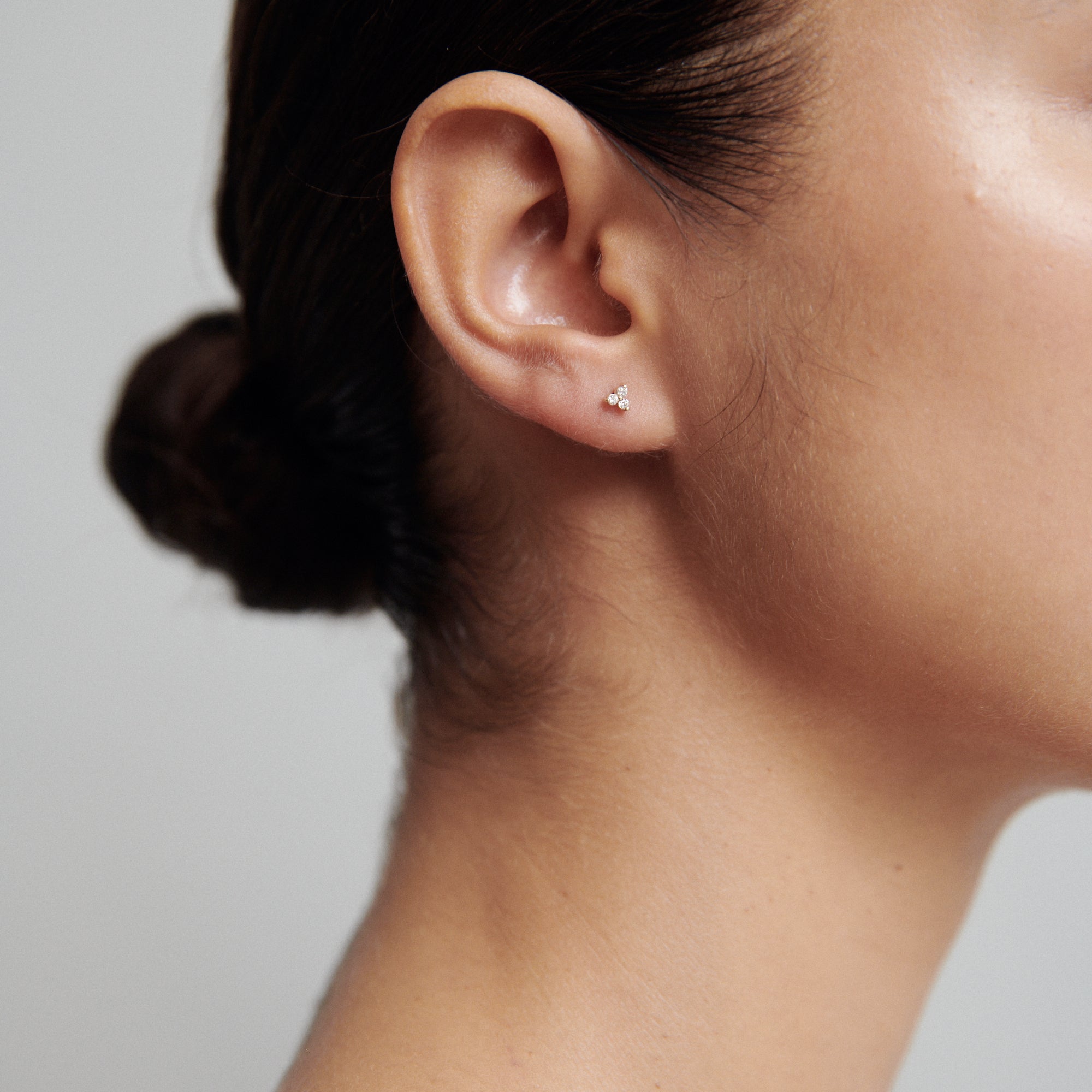 Woman's ear with a small diamond stud earring, hair styled in a neat bun, against a neutral background; luxury jewellery.