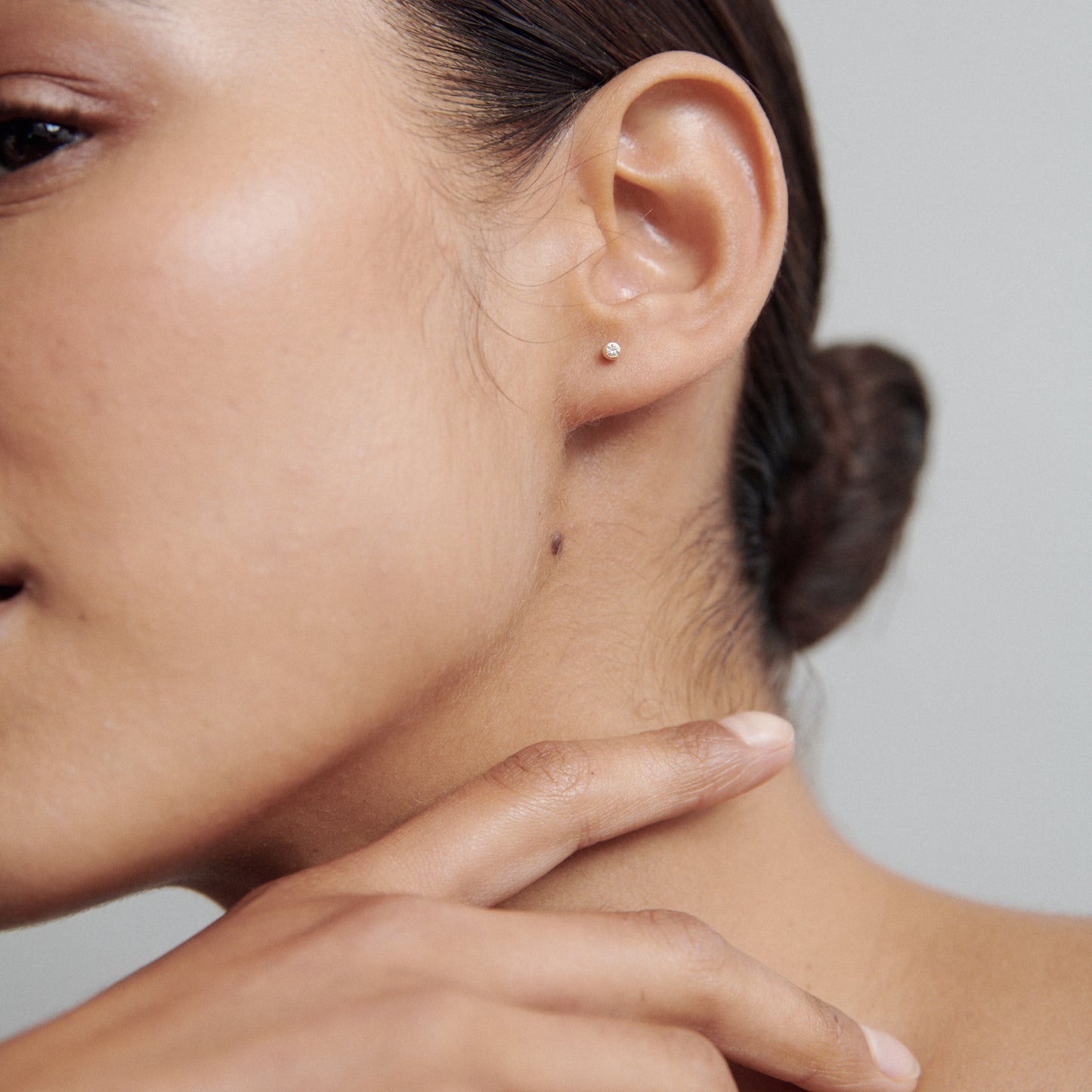 Close-up of a woman's ear wearing a small pearl stud earring. Her hand gently touches her neck. Elegant, minimalist jewellery style.