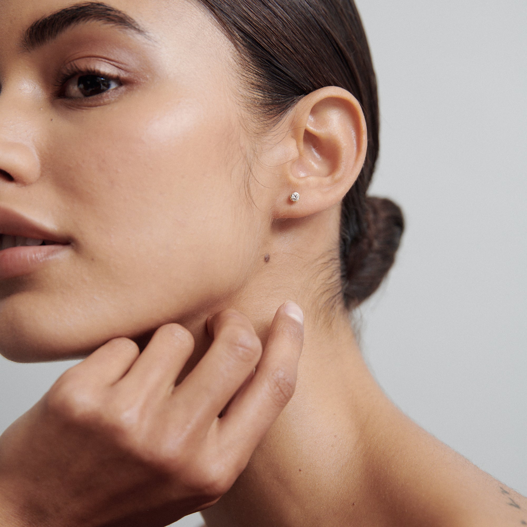 Close-up of a woman's face with a single diamond stud earring, smooth skin, and hair tied back neatly, touching her neck.