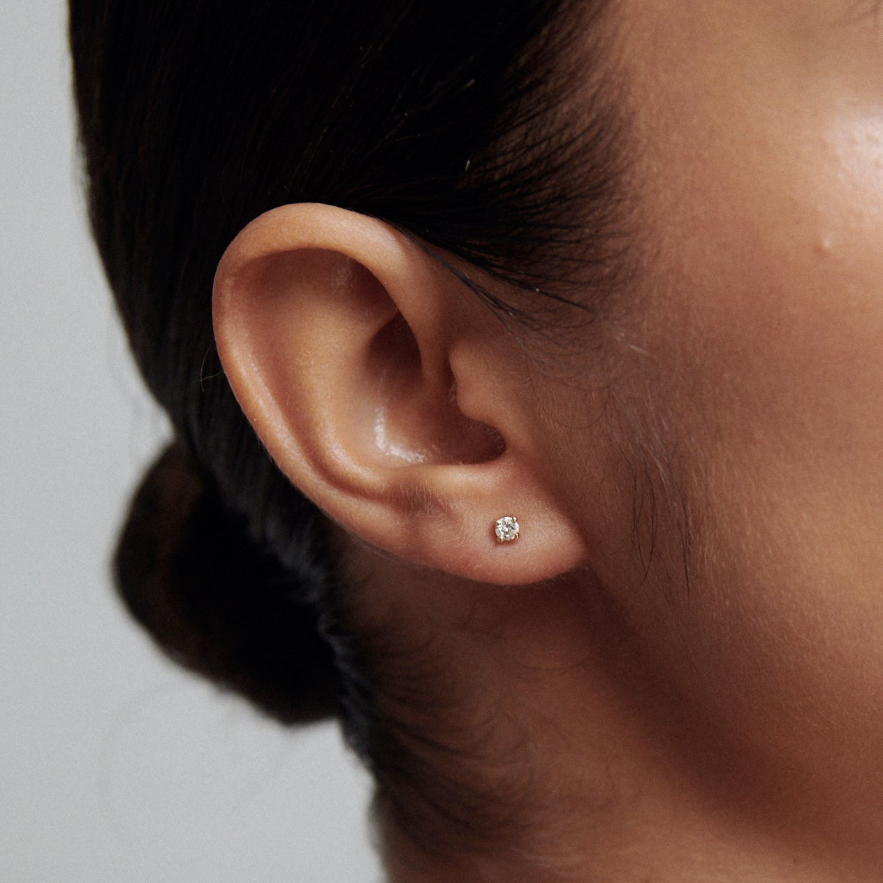 Close-up of a woman wearing a single small diamond stud earring. Her hair is neatly pulled back, showcasing the jewellery elegantly.