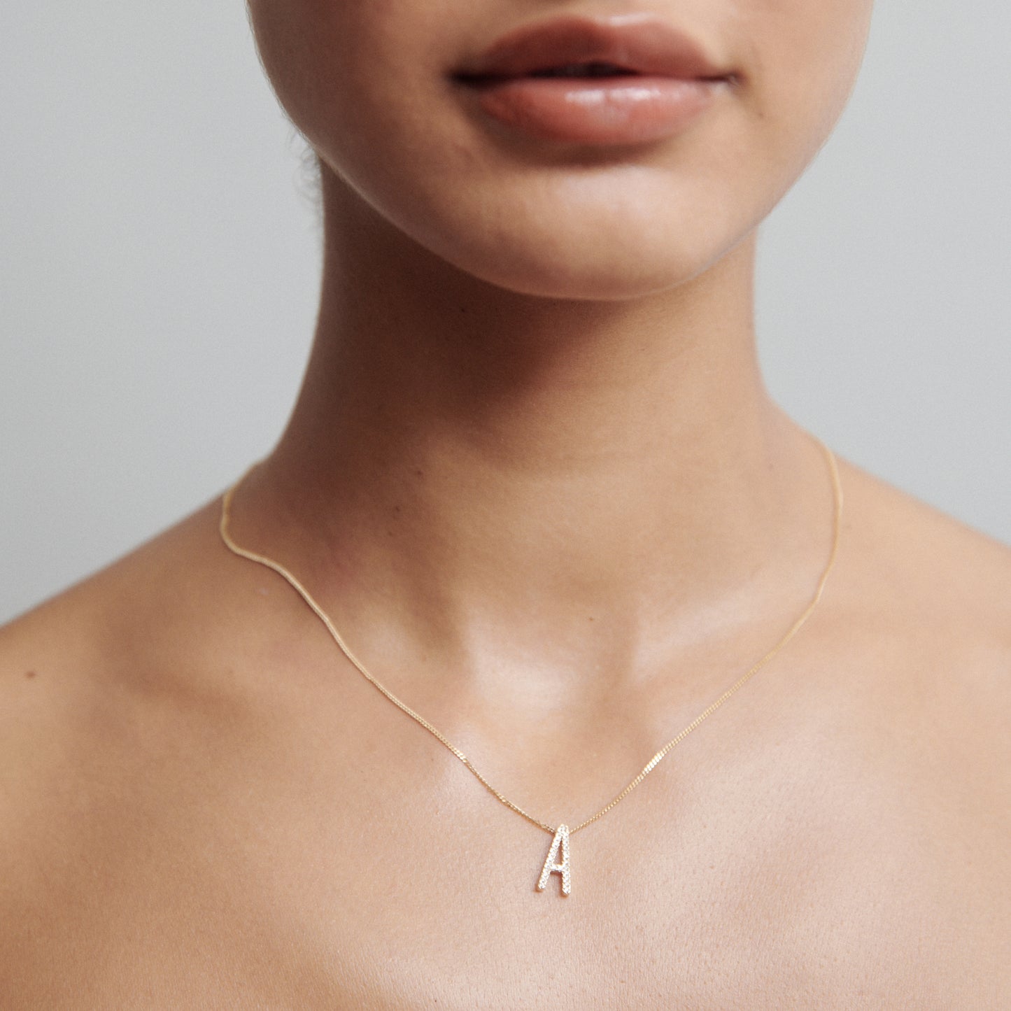 Woman wearing a delicate gold necklace featuring a diamond-studded letter "A" pendant, against a neutral background.