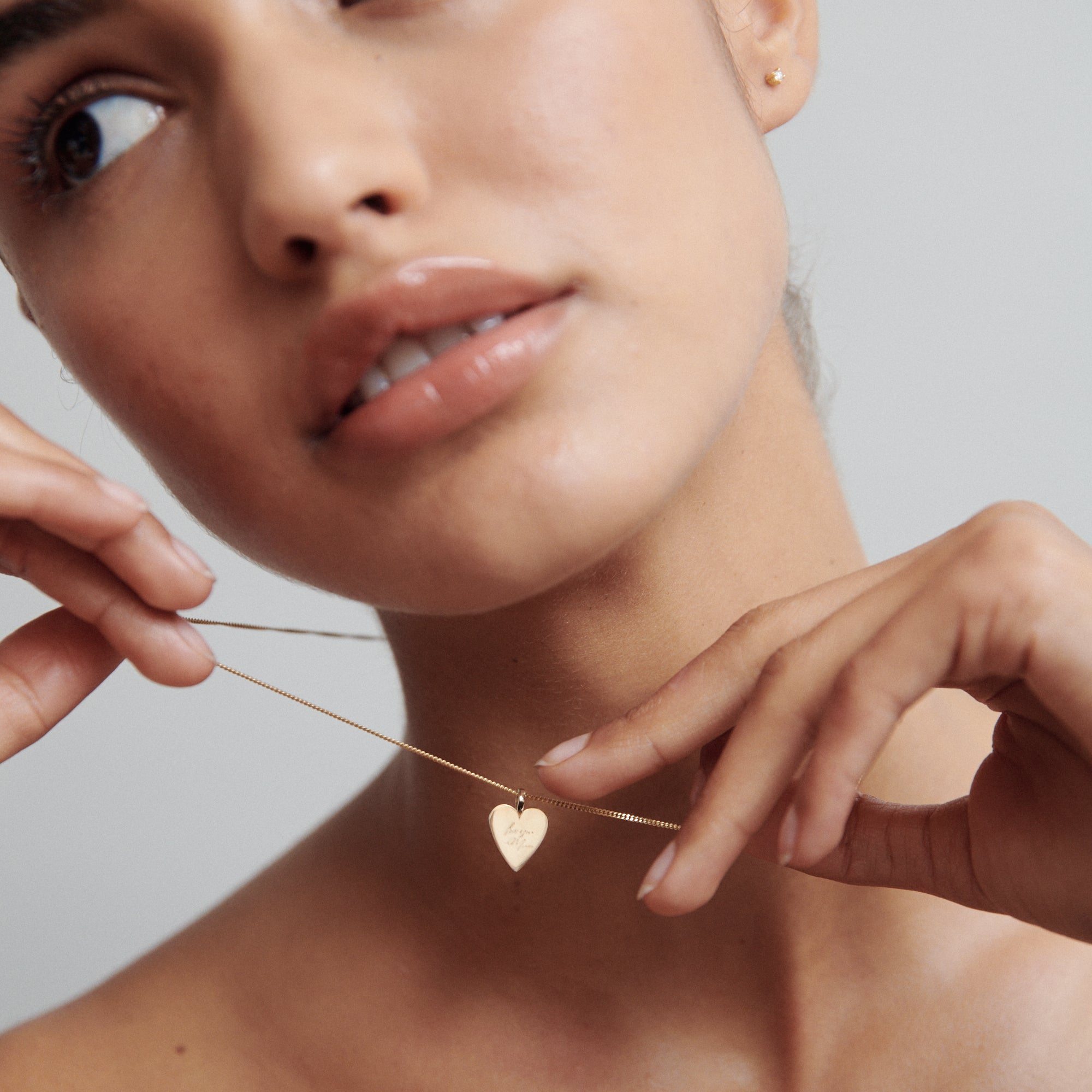 Close-up of person holding a gold heart necklace near their neck against a neutral background. Minimalist luxury jewellery style.