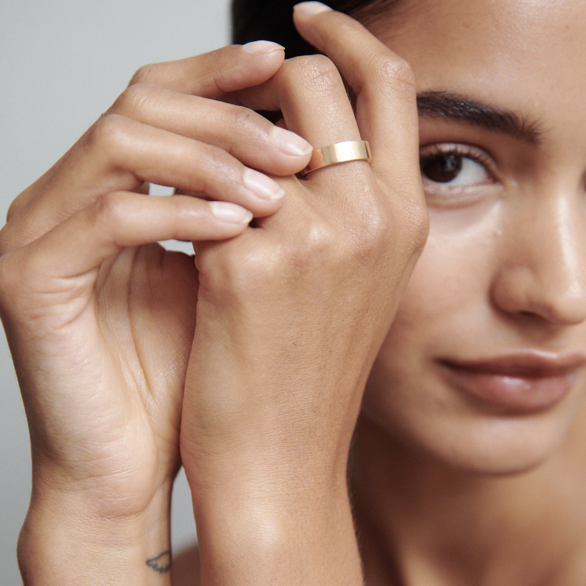 Woman displaying an elegant gold ring on her finger, showcasing luxury jewellery. Her hand is gently touching her face, highlighting the ring's sleek design.