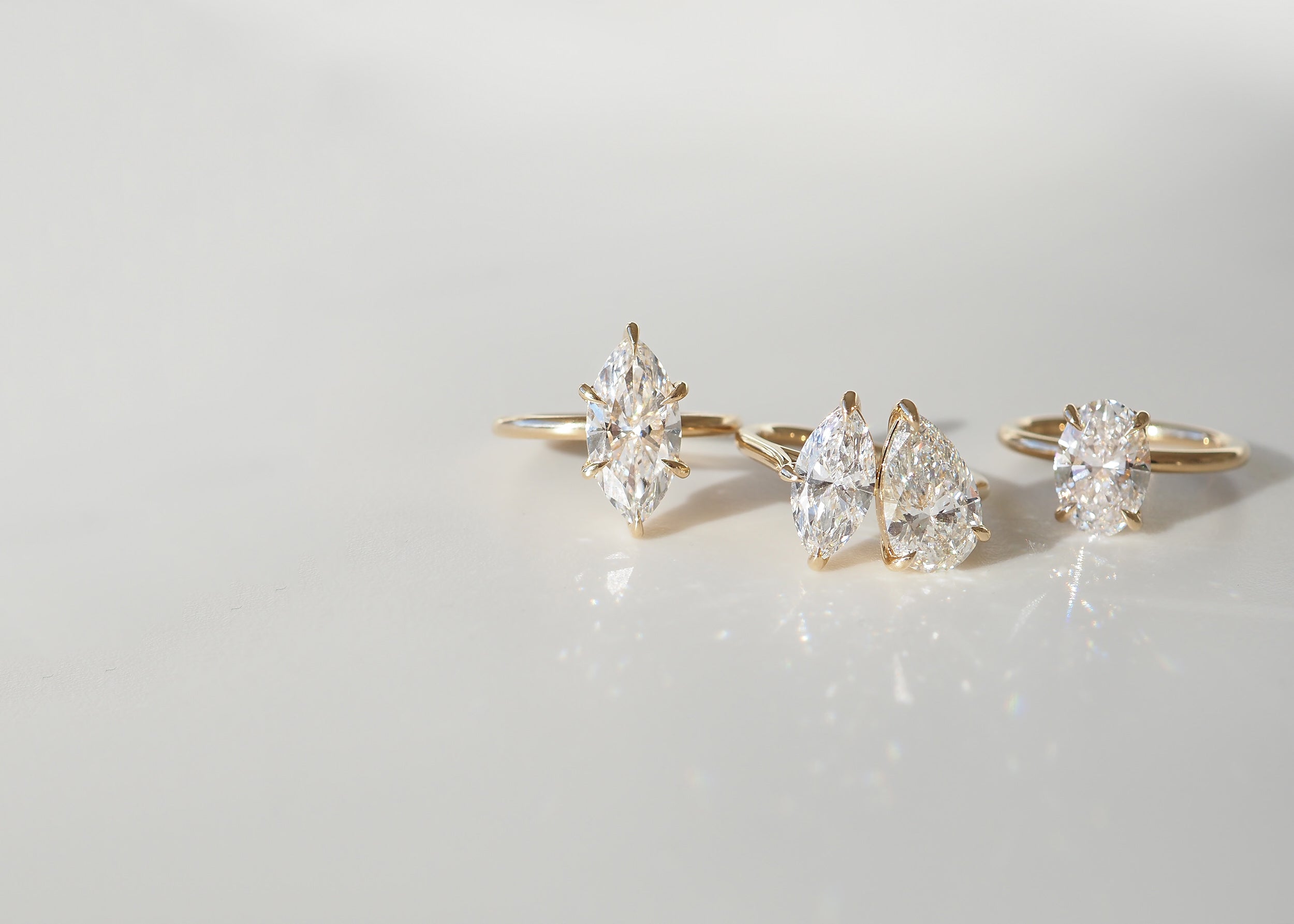 Three gold rings each showcase a unique diamond cut: marquise, pear, and round, elegantly placed on a smooth, reflective surface.