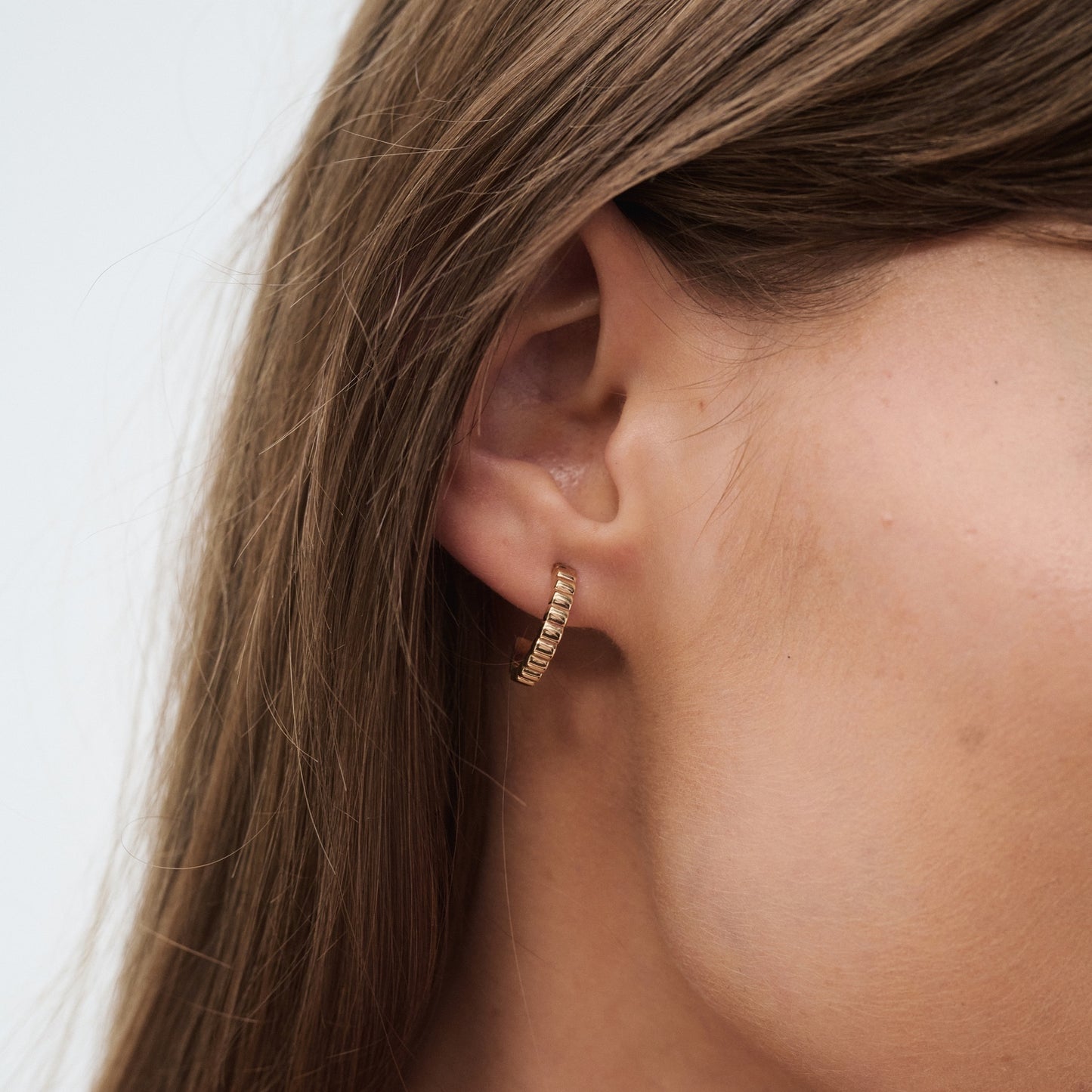 Close-up of a woman's ear adorned with a gold hoop earring featuring a row of small diamonds, showcasing luxury jewellery detail against her long brown hair.