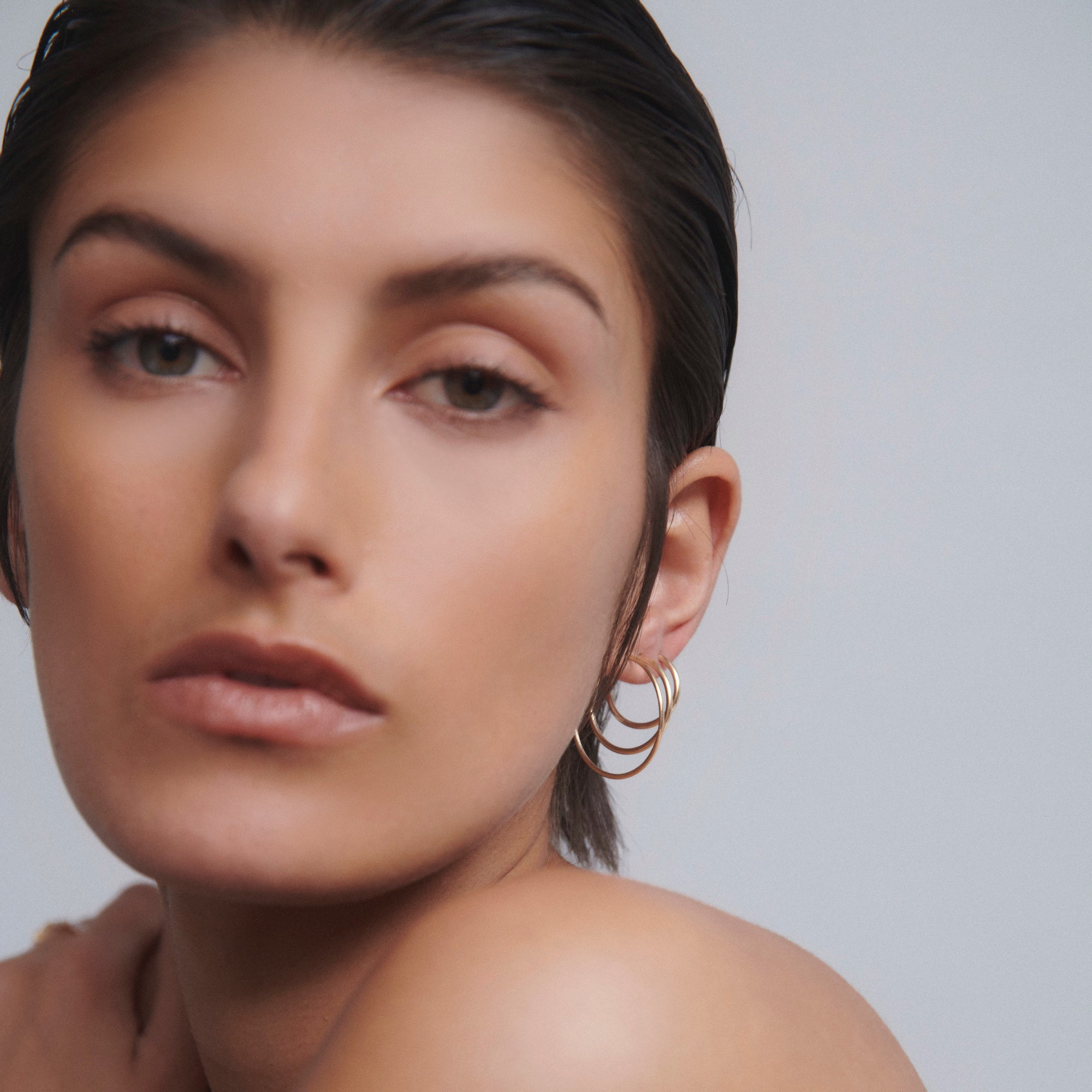 Woman with sleek hair wearing elegant gold hoop earrings, gazing softly at the camera. Minimalist style, luxury jewellery focus.