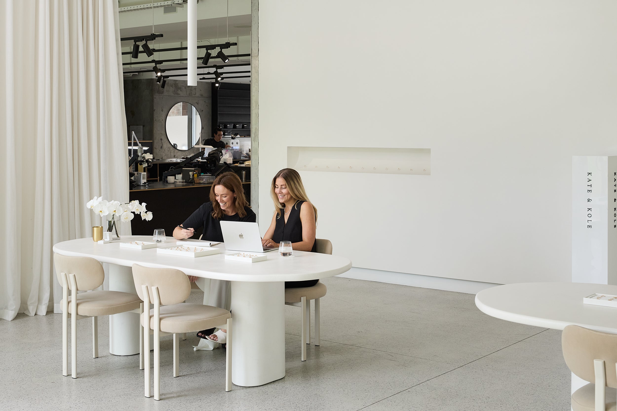 Two women sit at a white table with a laptop in a luxury jewellery store, featuring minimalist decor, white chairs, and elegant lighting.