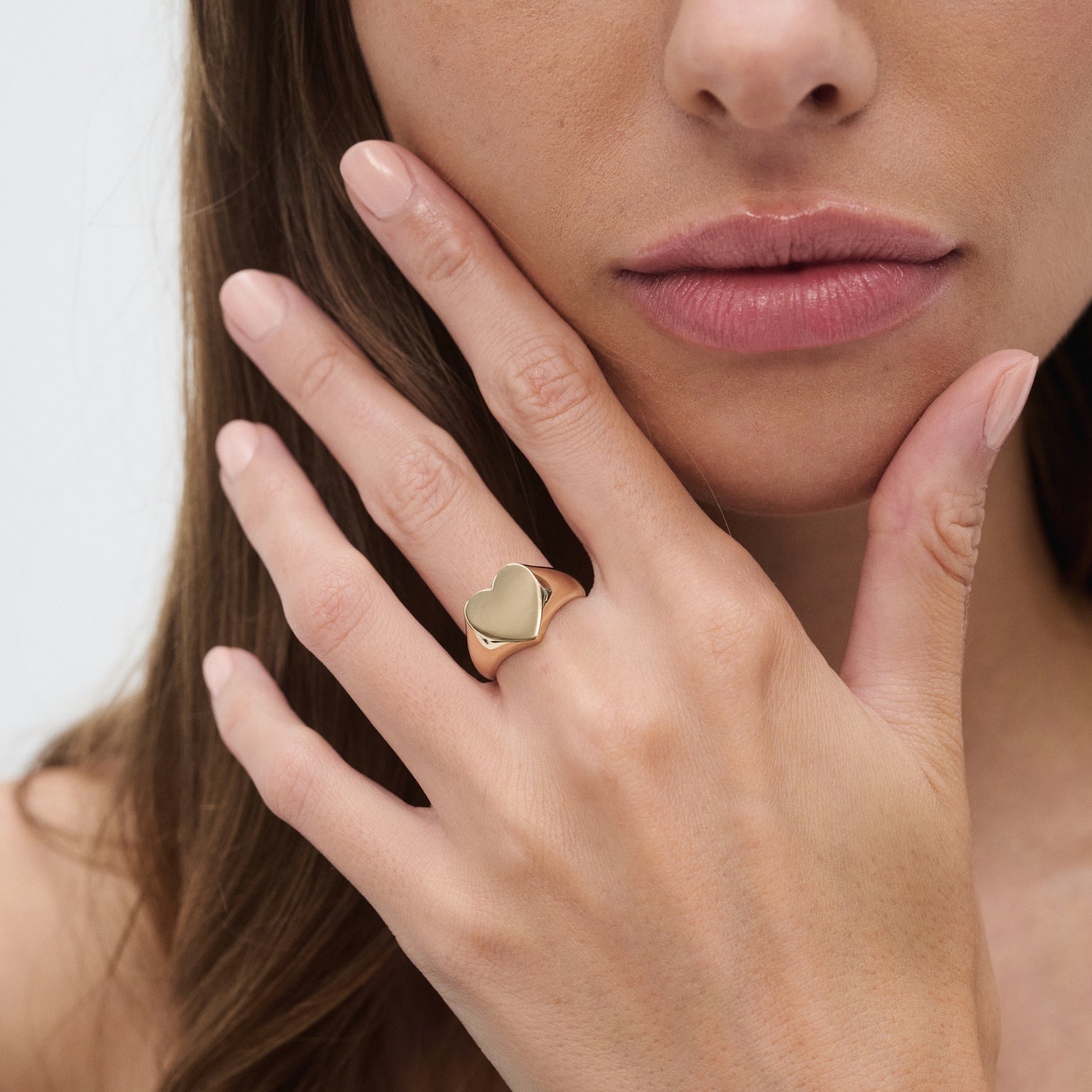 Woman's hand with a gold heart-shaped ring on her finger, touching her face. Luxury jewellery, elegance, minimalist style.