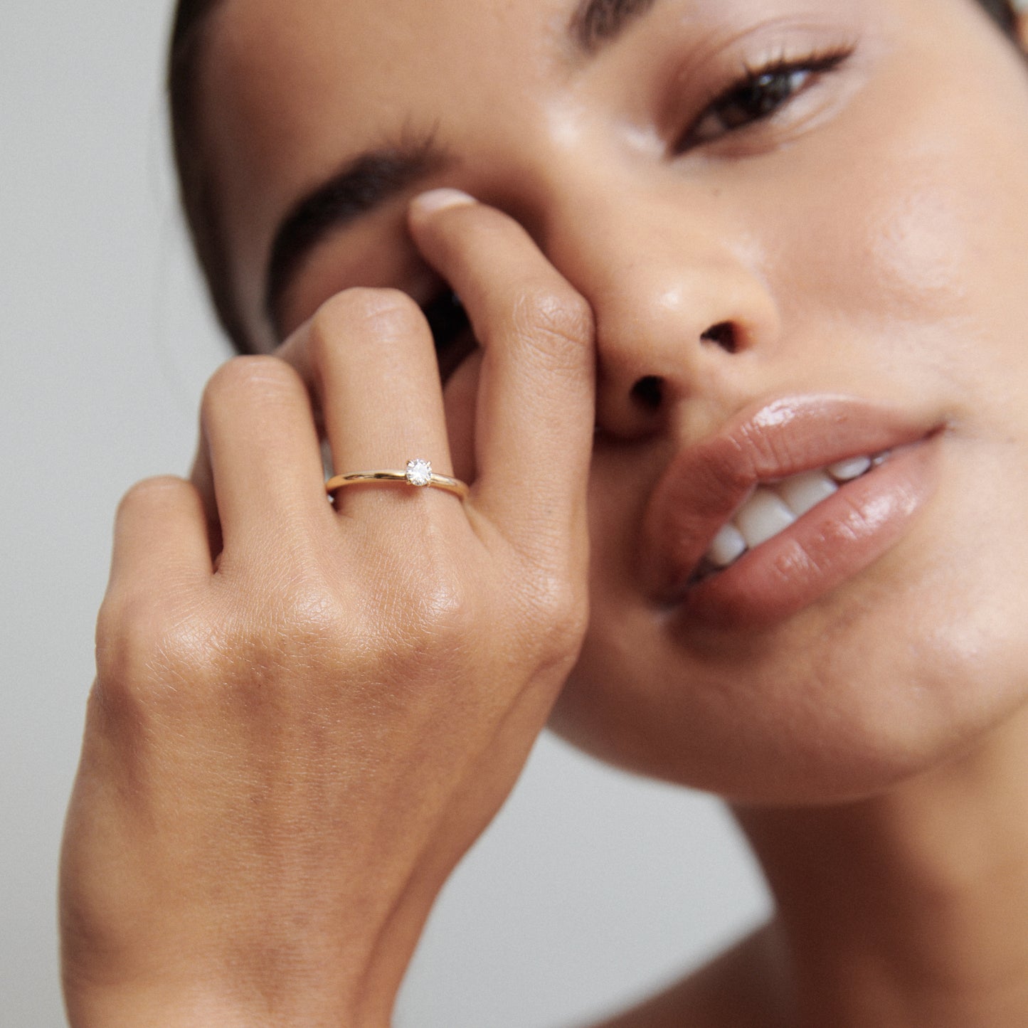 Woman shows a gold ring with a solitaire diamond on her finger, highlighting luxury jewellery. She has a serene expression with natural makeup.