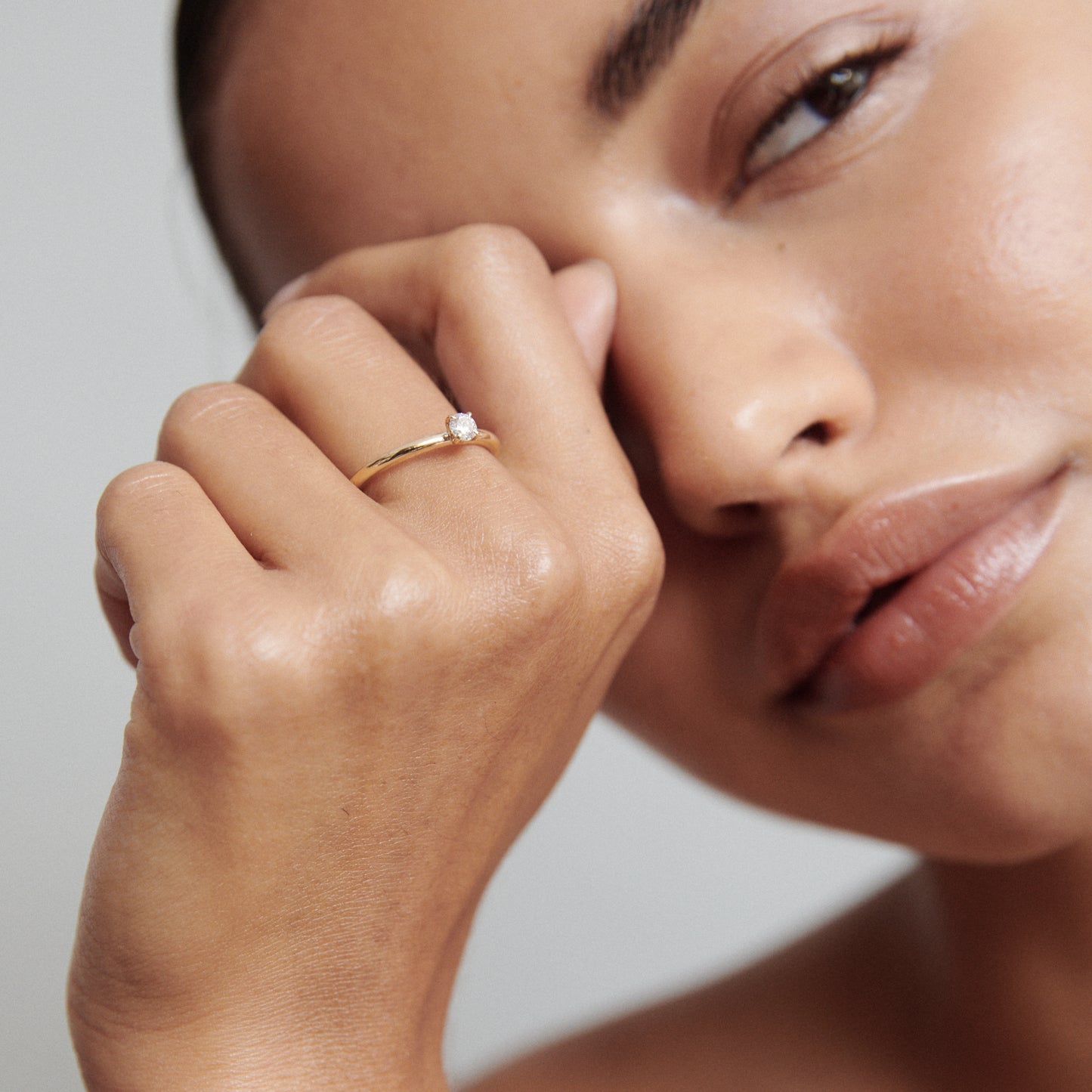 Woman showcasing a gold engagement ring with a solitaire diamond, resting near her face against a neutral backdrop. Luxury jewellery close-up.