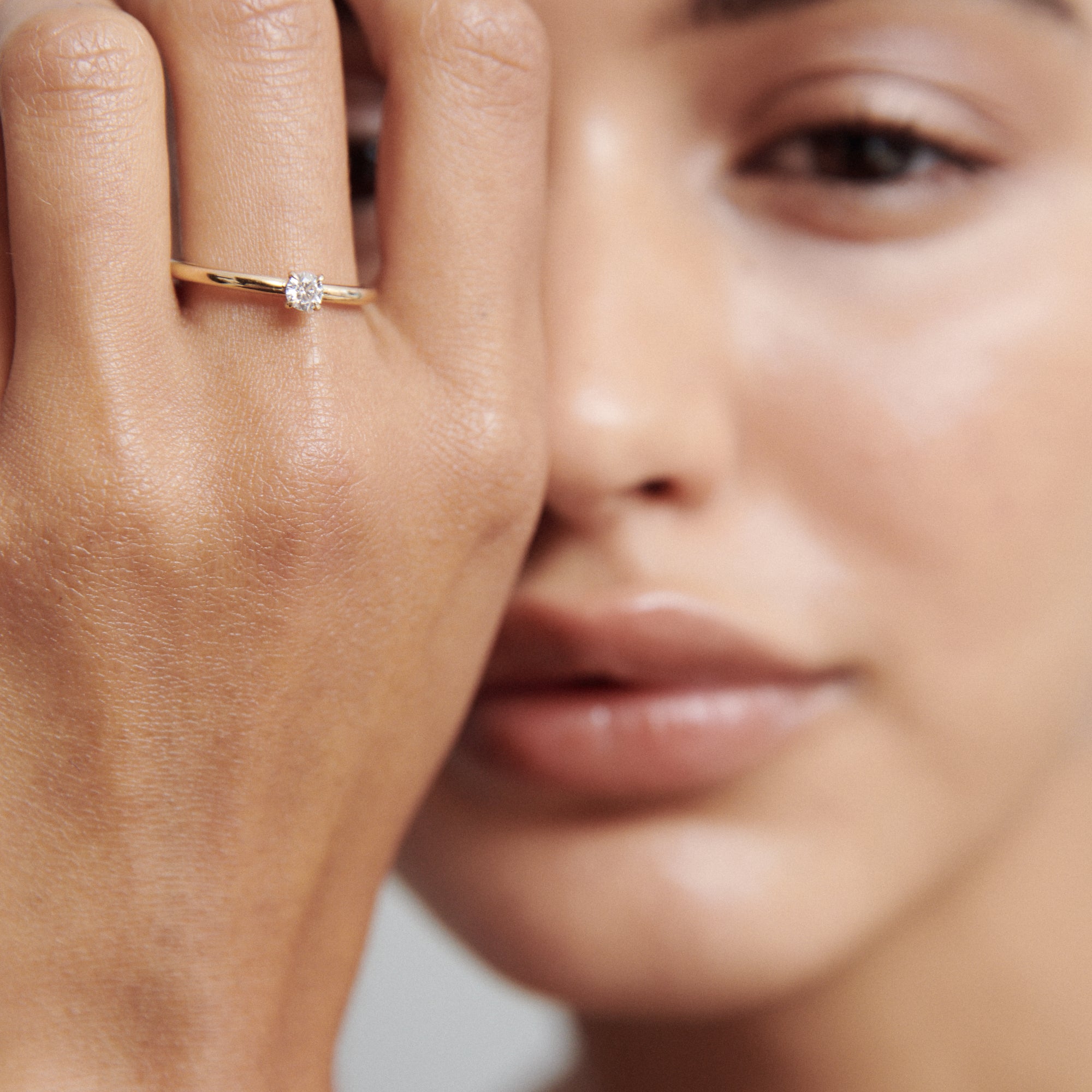 Close-up of a woman's hand with a delicate gold ring featuring a single round diamond, her face softly blurred in the background. Luxury jewellery.