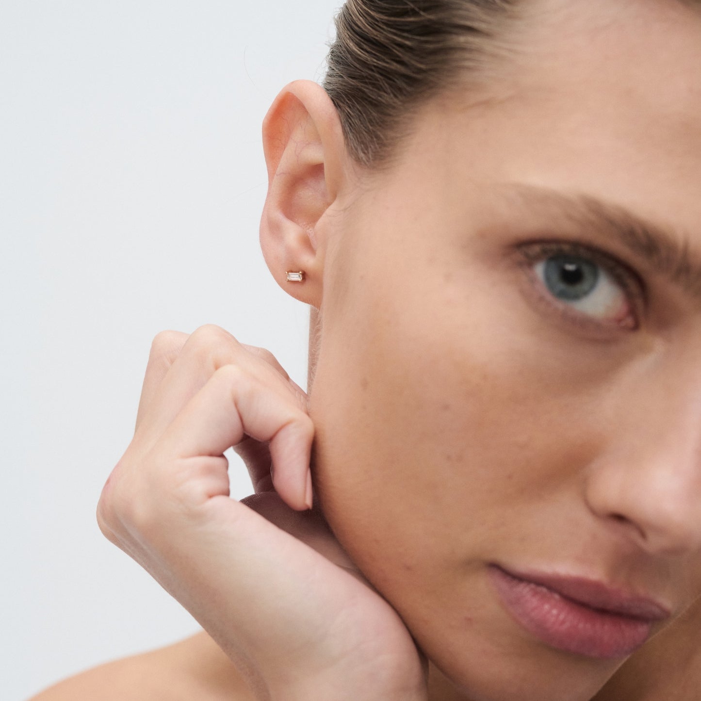 Close-up of a woman's face showcasing a single diamond stud earring in her left ear, with a neutral background.