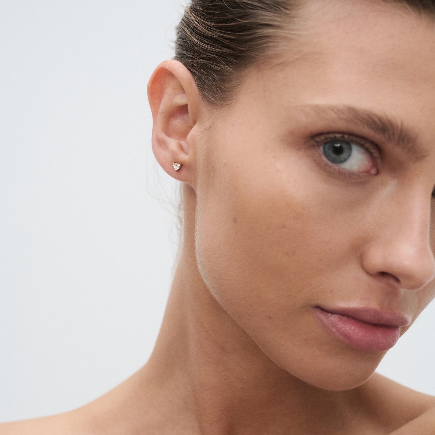 Close-up of a woman wearing a small, elegant diamond stud earring. Her hair is tied back, highlighting the jewellery against clear skin.
