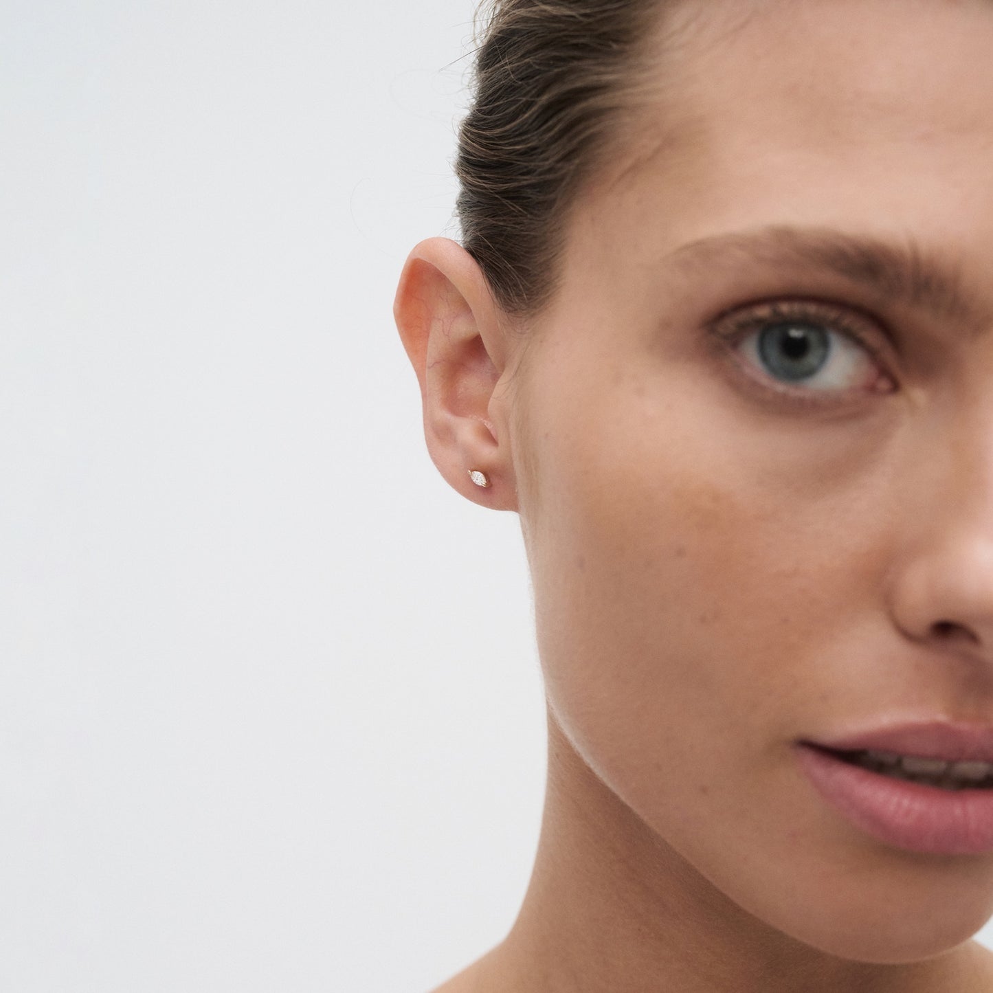 Close-up of a woman's face wearing a minimalist gold stud earring, highlighting luxury jewellery on smooth skin with a neutral expression.