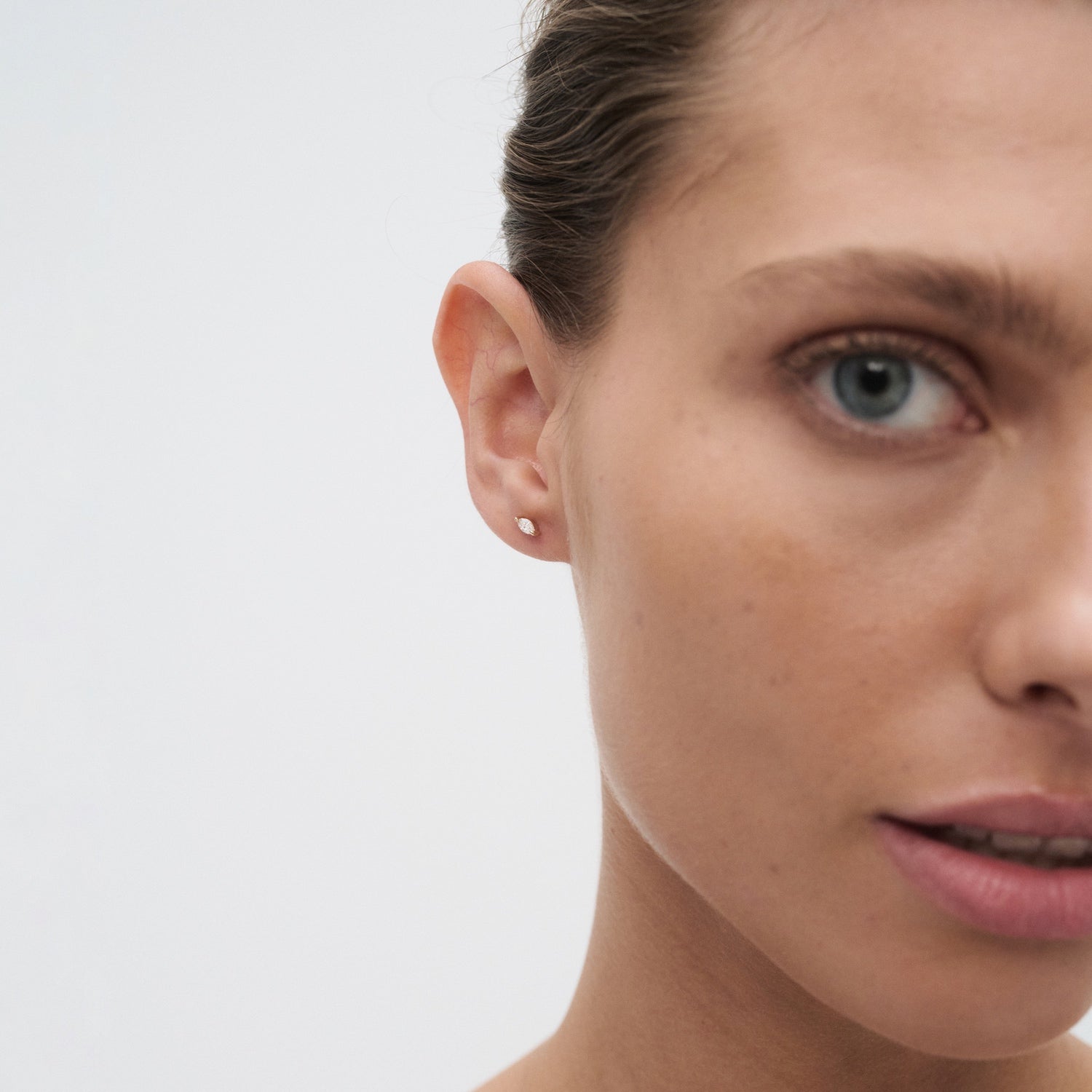 Close-up of a woman's face wearing a minimalist gold stud earring, highlighting luxury jewellery on smooth skin with a neutral expression.