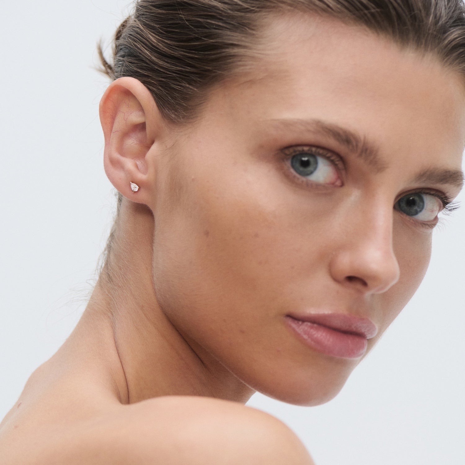 Woman with sleek hair wearing a single diamond stud earring, gazing sideways. Luxurious jewellery fashion. Minimalist elegance.
