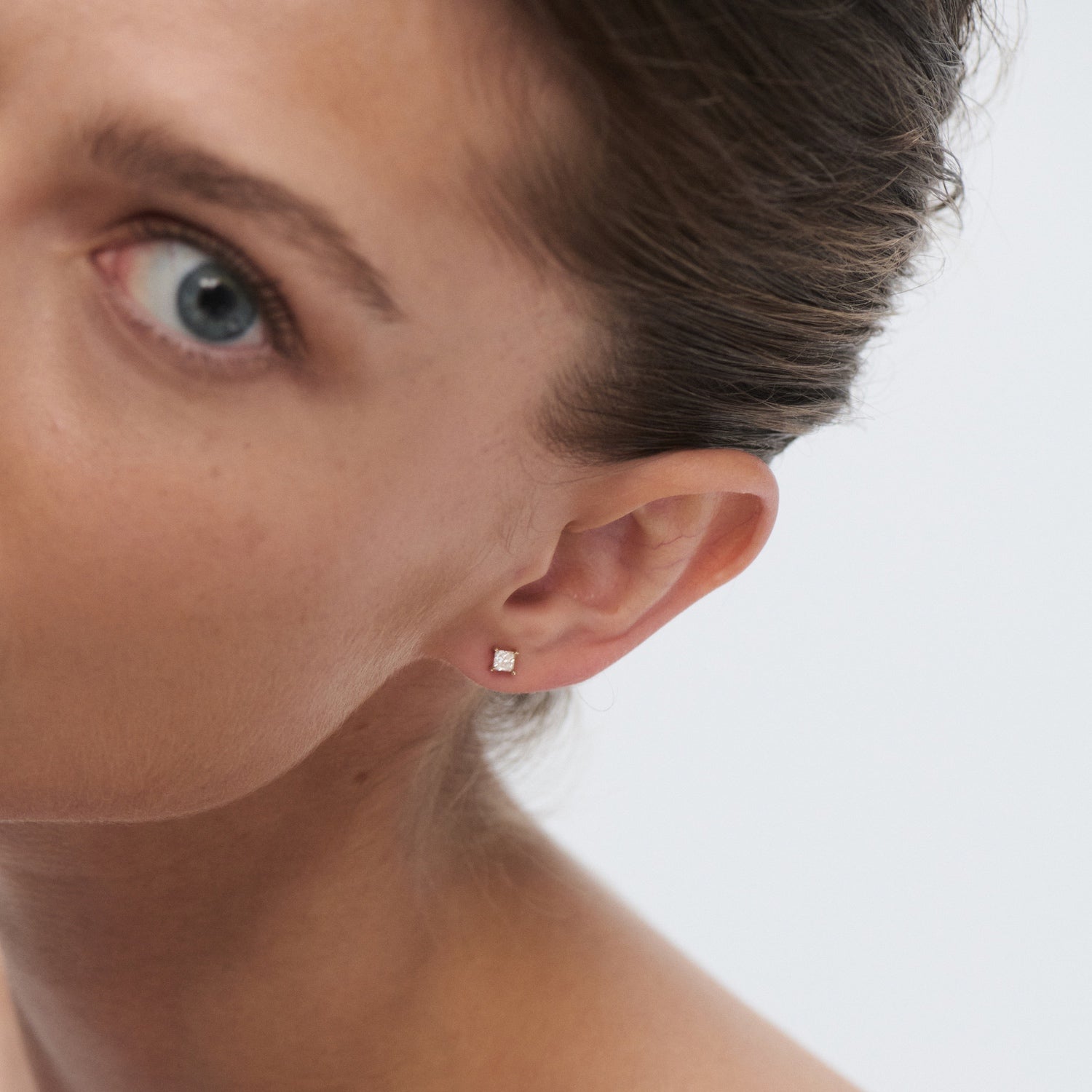 Close-up of a woman's ear wearing an elegant stud earring with a square diamond set in gold. Luxurious, minimalist jewellery design.