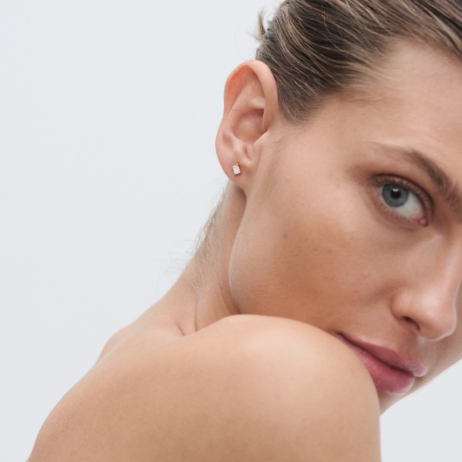 Woman with slicked-back hair wearing a single diamond stud earring on the left ear, against a plain background. Luxurious, elegant jewellery.