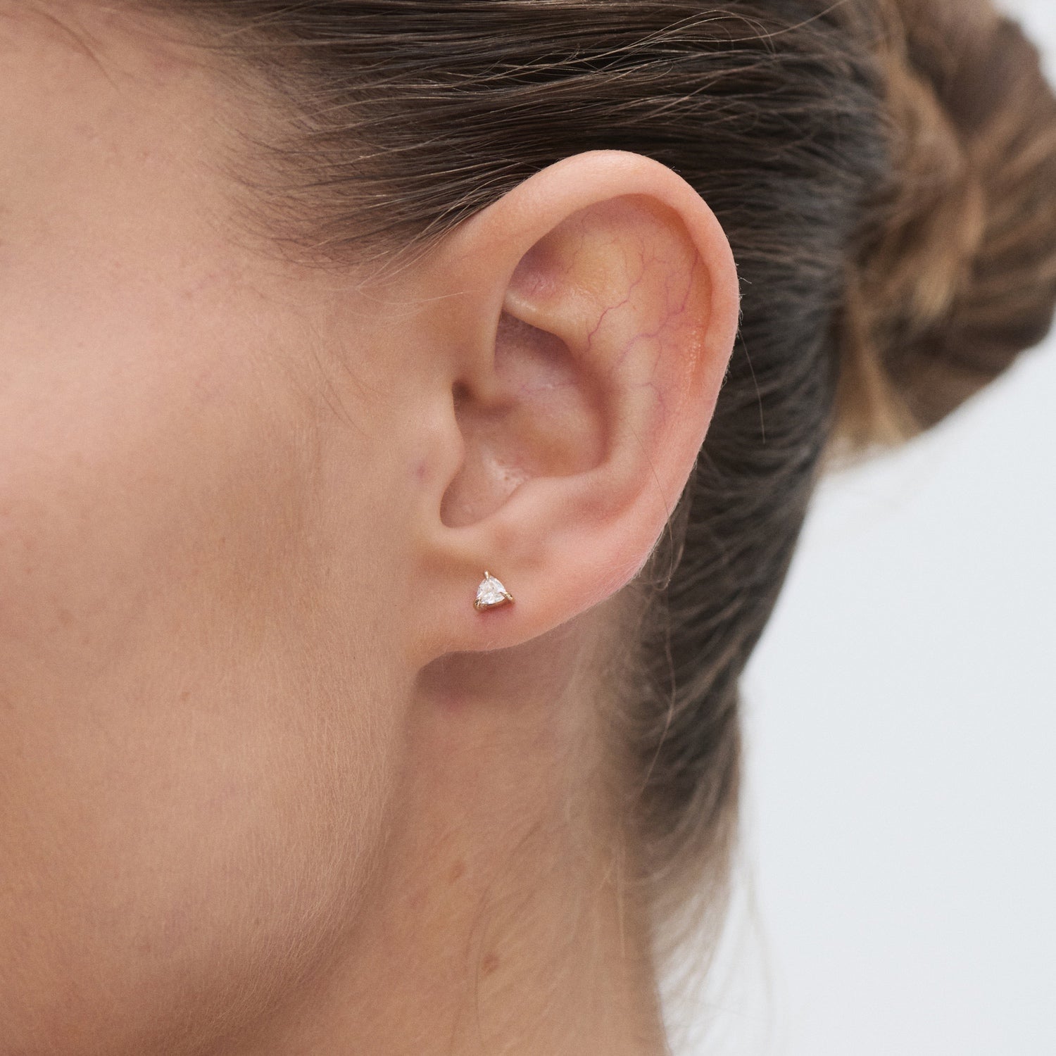Close-up of a woman wearing a single triangular diamond stud earring against a light background. Her hair is tied in a neat bun.