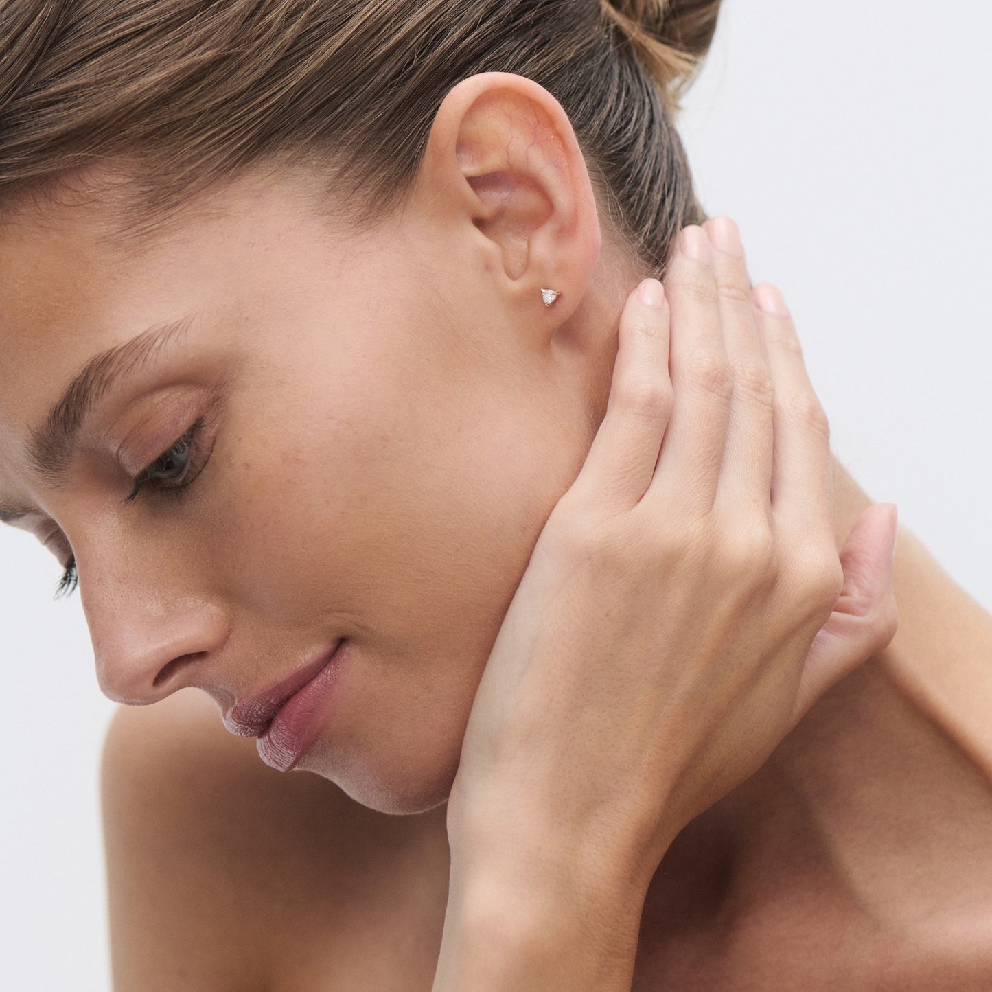 Woman with elegant hair bun wearing a minimalist gold stud earring, gently touching her ear. Luxury jewellery focus, serene expression.