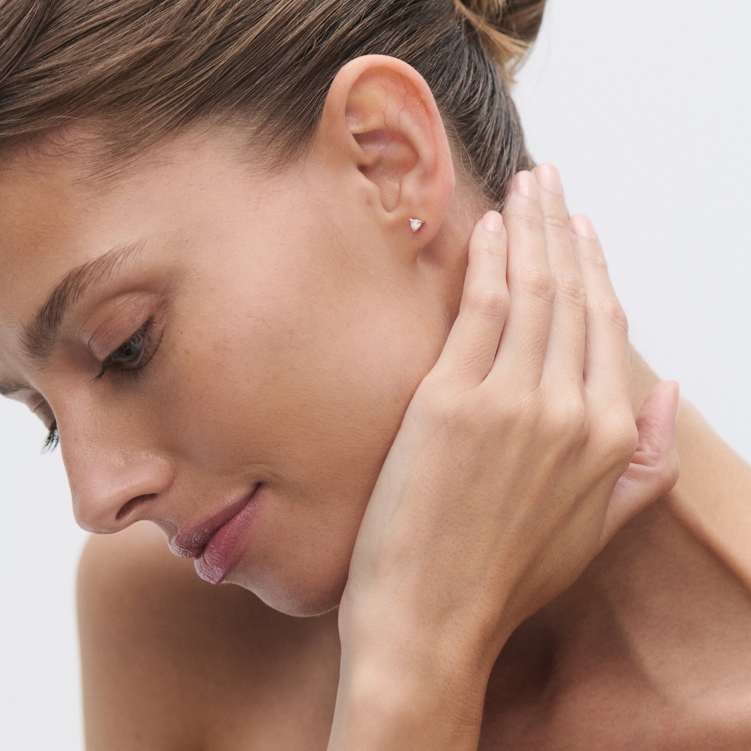 Woman with elegant hair bun wearing a minimalist gold stud earring, gently touching her ear. Luxury jewellery focus, serene expression.