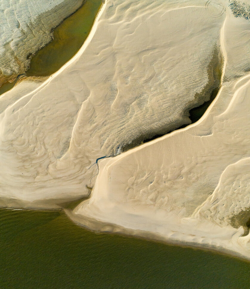 Aerial view of a serene sandy beach with water inlets, rippled textures, and calm greenish waves, evoking coastal luxury and tranquility.
