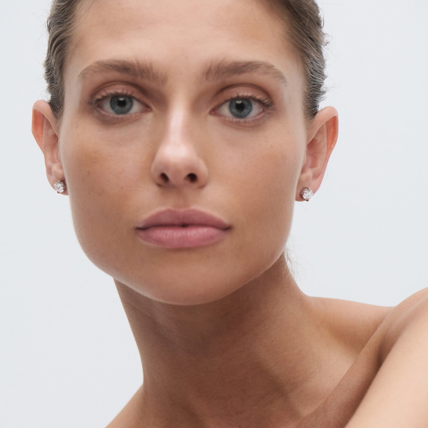 Woman wearing elegant diamond stud earrings, neutral makeup, and hair pulled back, against a plain grey background. Luxurious, high-end jewellery.