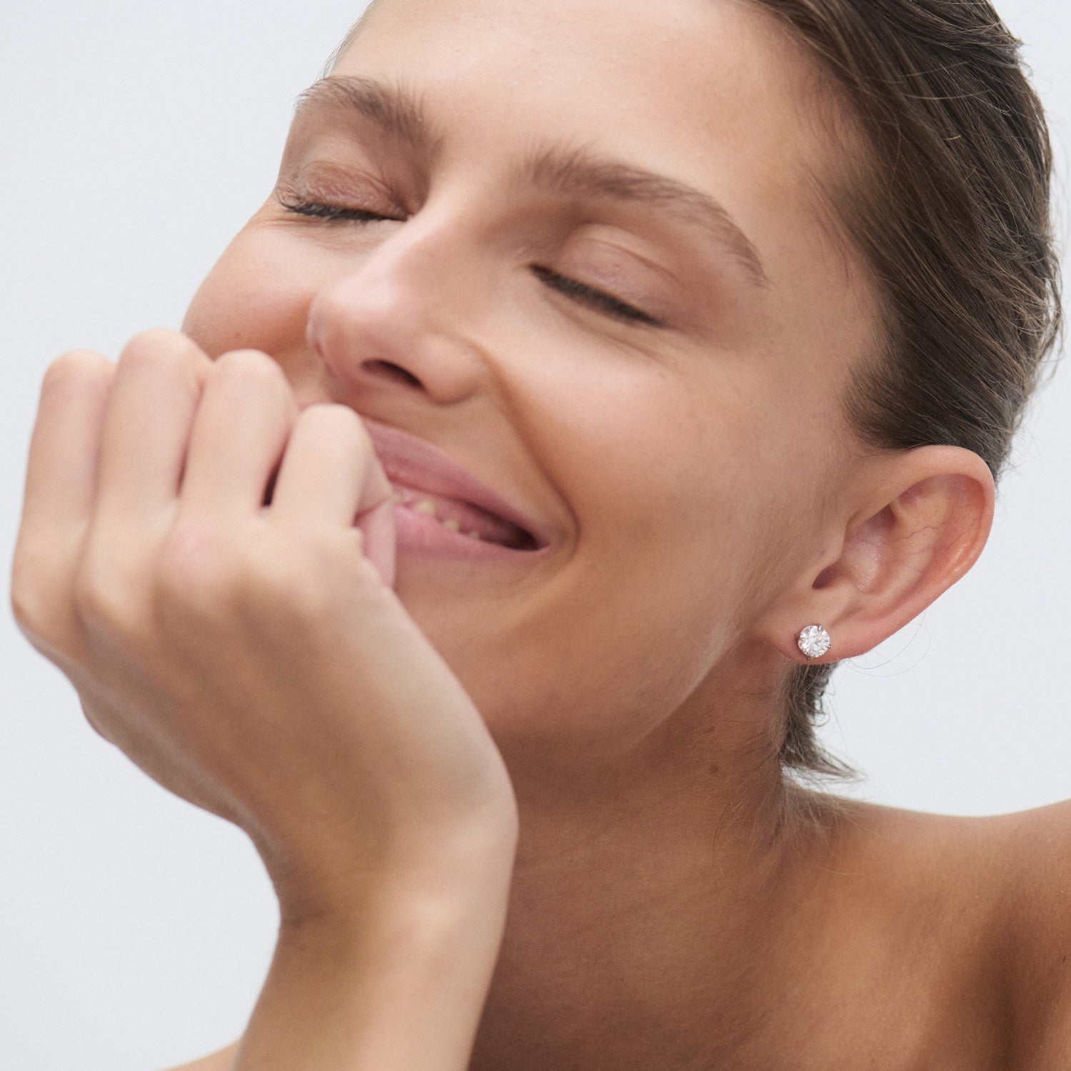 Smiling woman wearing elegant diamond stud earrings, eyes closed, showcasing luxury jewellery in soft lighting.