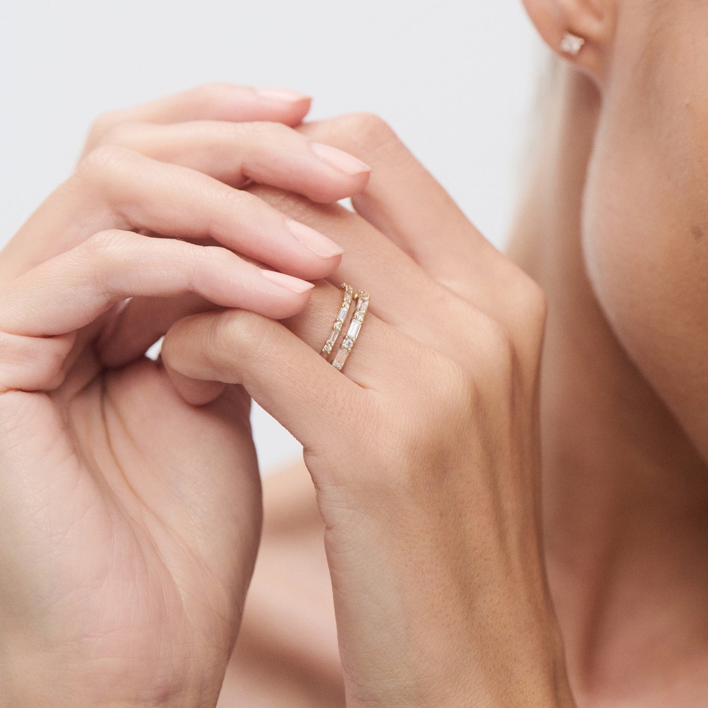 Elegant woman wearing stacked diamond rings on left hand, touching face. Luxury jewellery with gold bands and clear gemstones. Minimalist style.