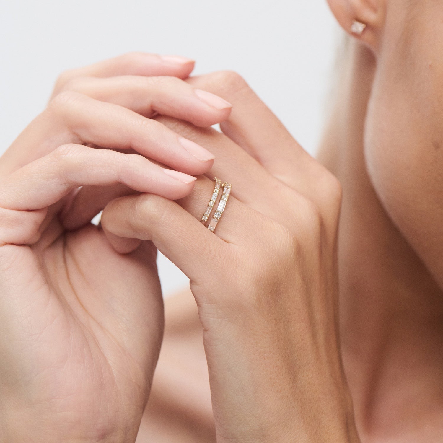 Elegant woman wearing stacked diamond rings on left hand, touching face. Luxury jewellery with gold bands and clear gemstones. Minimalist style.