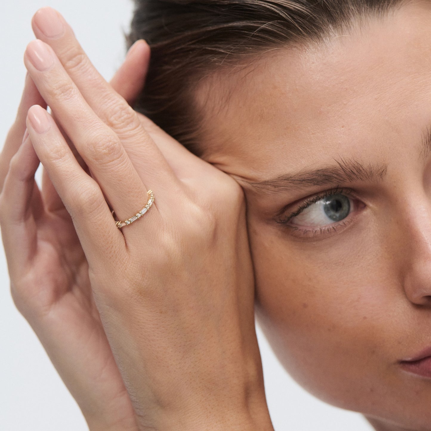 Close-up of a person holding their face, wearing a delicate gold band on their finger. Luxurious and minimalist jewellery style.