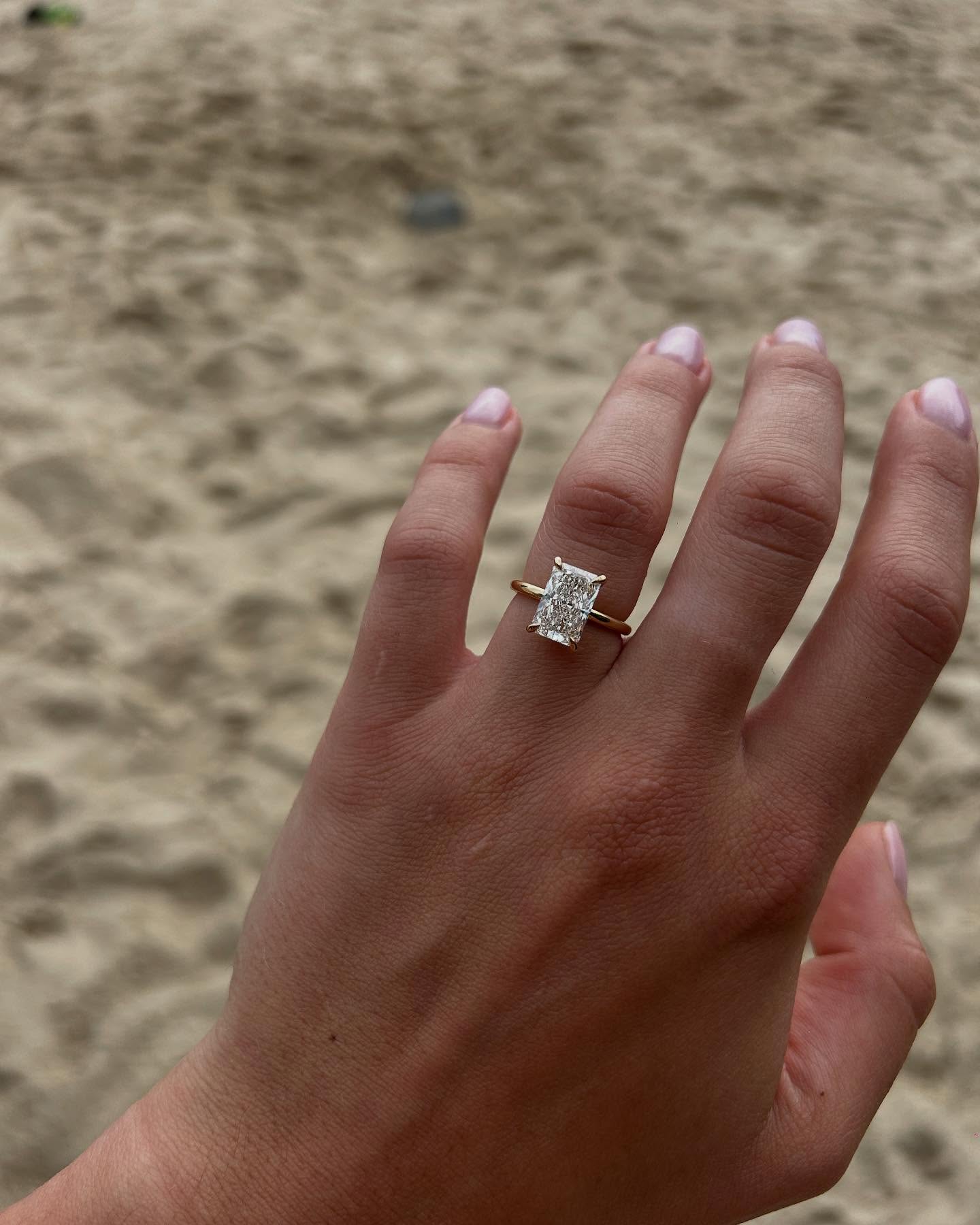 Hand displaying a rectangular-cut diamond ring with gold band against a sandy beach backdrop. Luxury jewellery elegance.